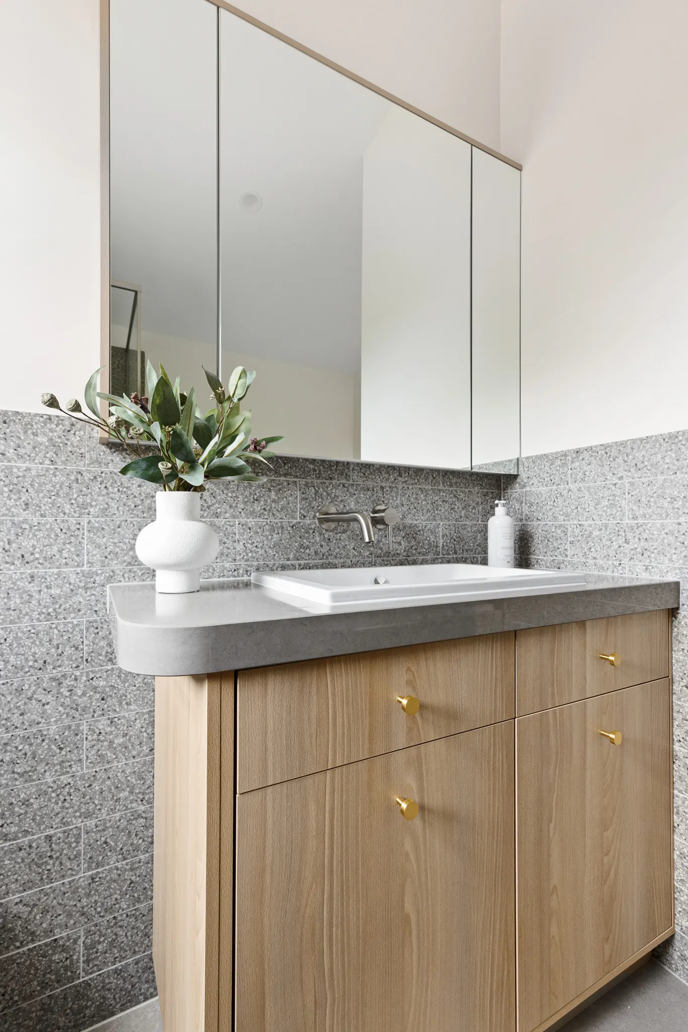 Timber vanity with brass knobs, curved stone benchtop and grey tiles below mirrored cabinets