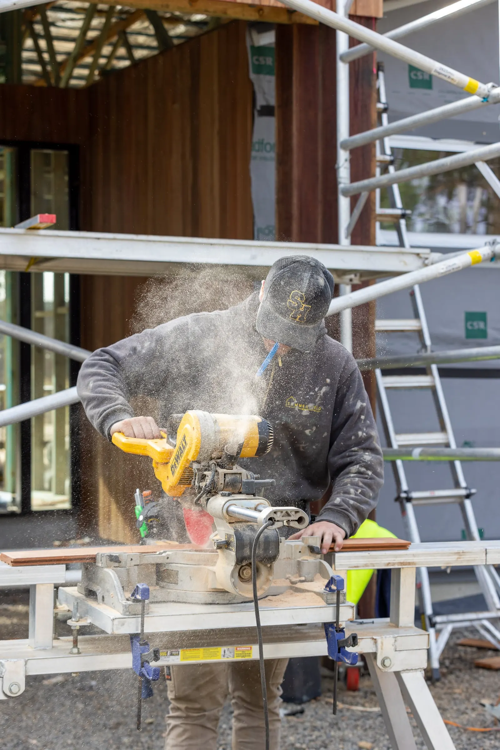 A Summerfield Homes carpenter cutting timber on a mitre saw, sawdust in the air, during the Dohoneys Rd build