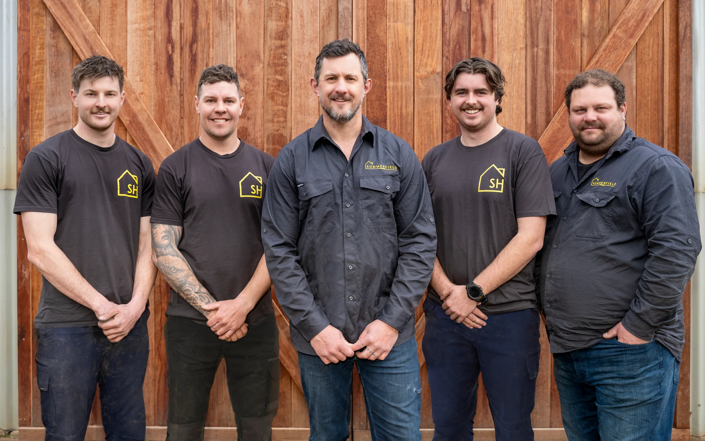 The Summerfield Homes crew of five in Summerfield workwear outside a timber barn door &mdash; Angus, Jesse, Casey, Conor and Hayden