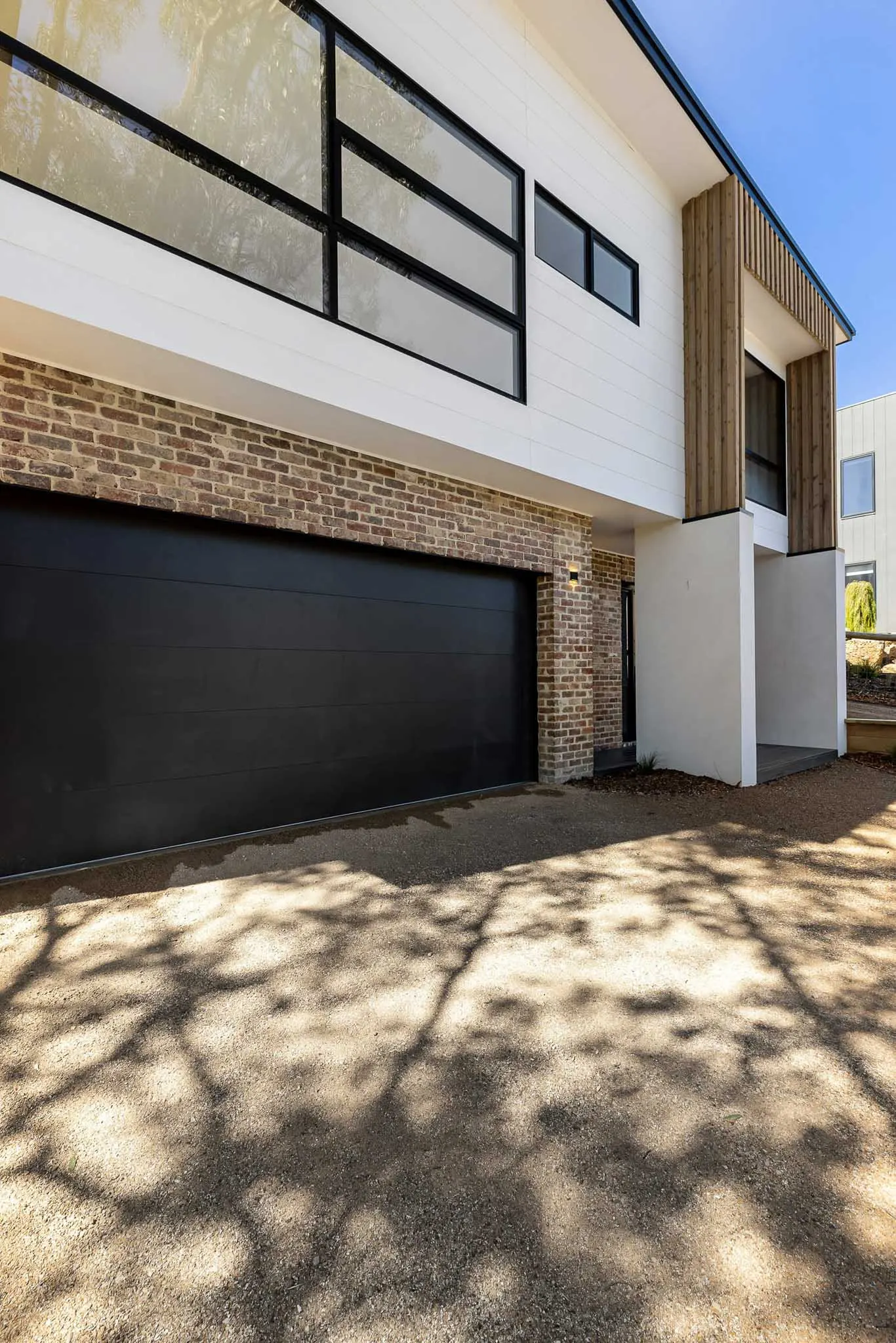Black sectional garage door in the brick lower storey, white cladding and battens above