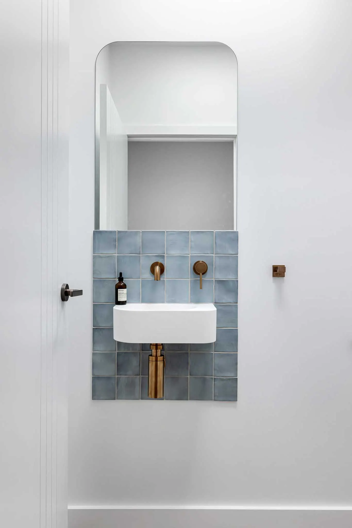 Wall-hung basin on a blue tile panel with brass tapware and exposed trap, arched mirror
