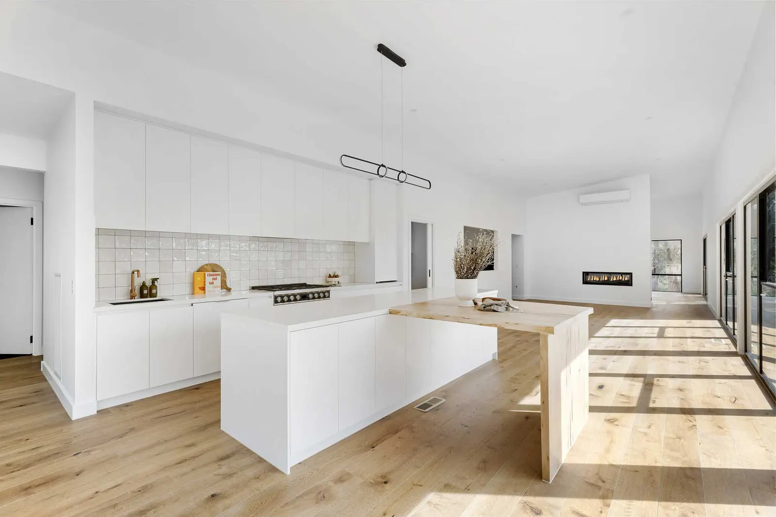 Open-plan kitchen and living under a raked ceiling, island with timber table and fireplace beyond