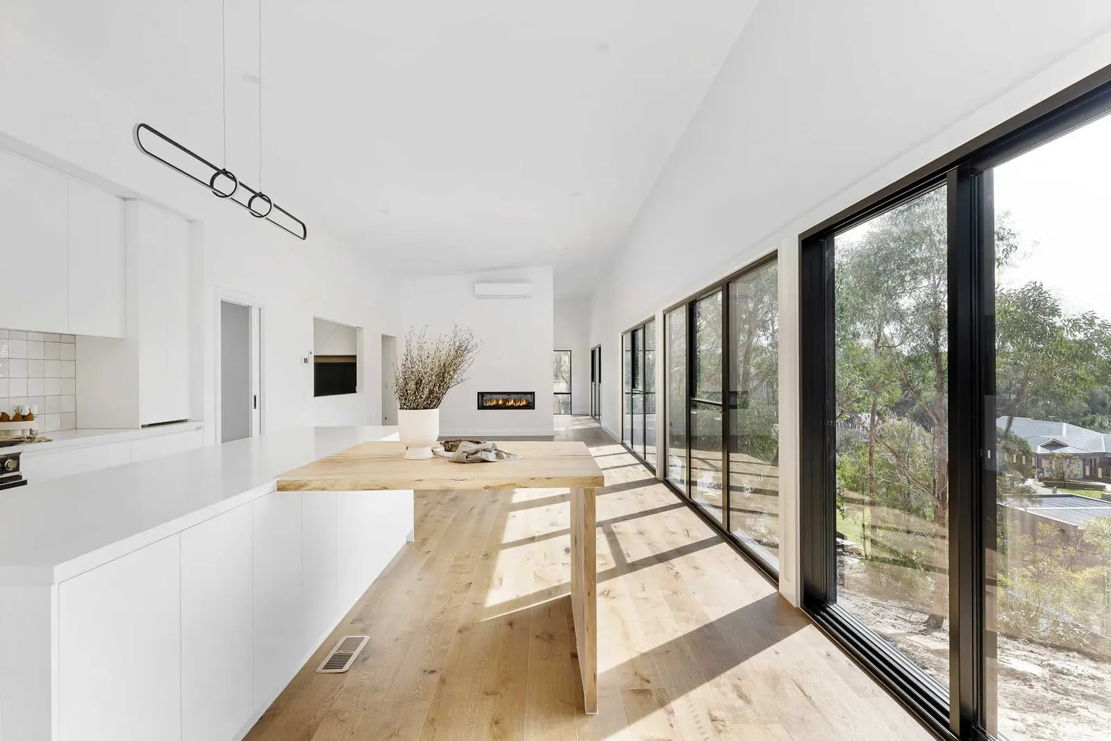 Timber table off the island beside full-height glazing to the bush, fireplace at the far end