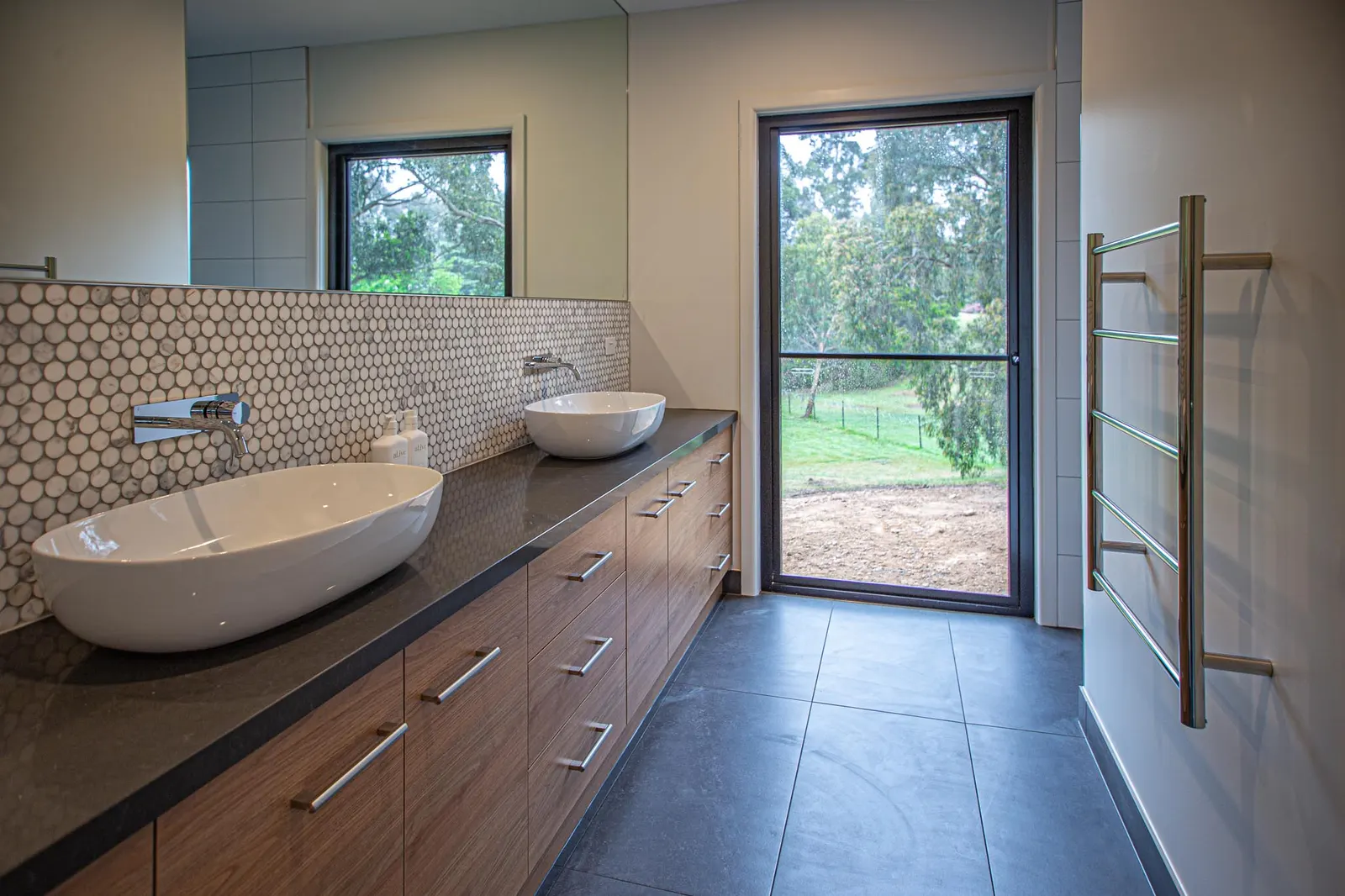 Twin oval vessel basins on a dark benchtop with timber joinery and a mosaic splashback