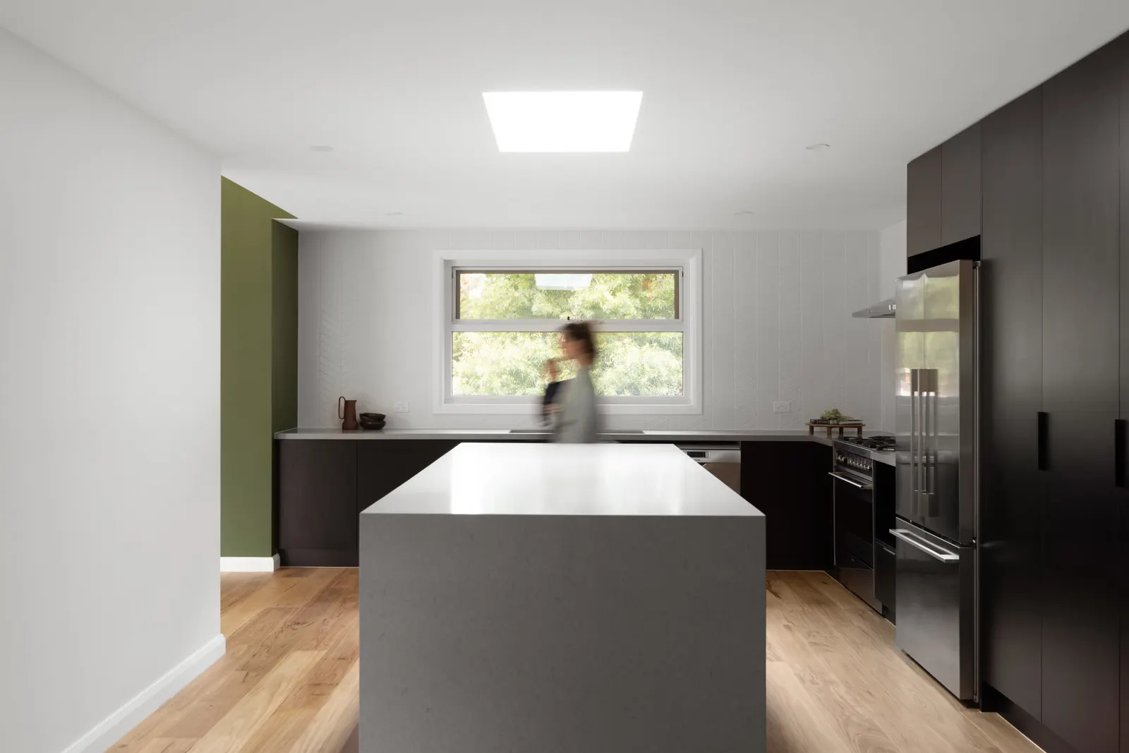 A figure moves past the kitchen island beneath the skylight, window over the sink