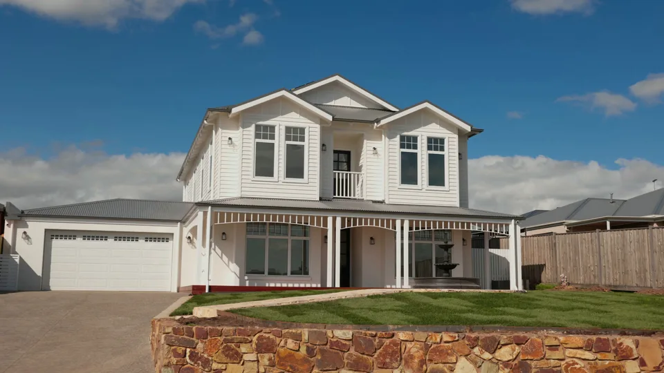 White two-storey weatherboard home with a verandah, above a stacked-stone wall at Mount Gisborne