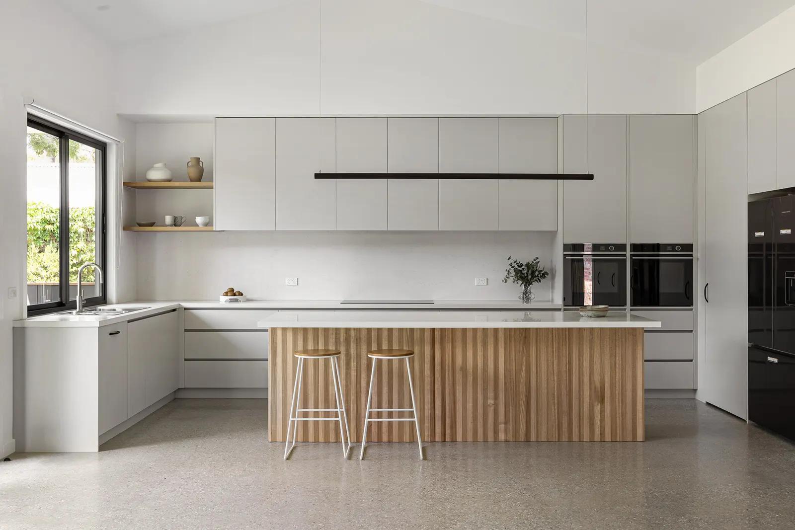 Kitchen with pale grey joinery, fluted timber island and two stools on polished concrete