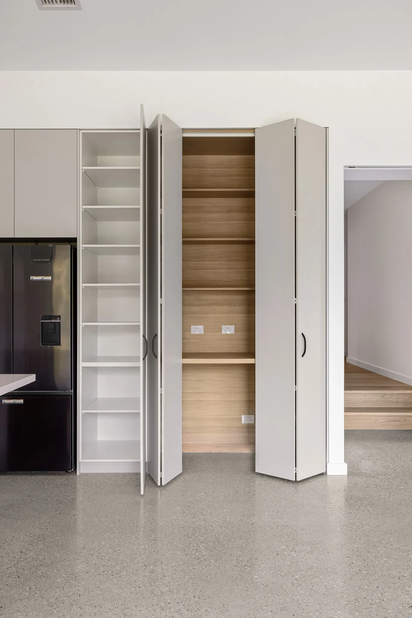 Bi-fold cupboard opened to timber-lined shelving beside a black fridge