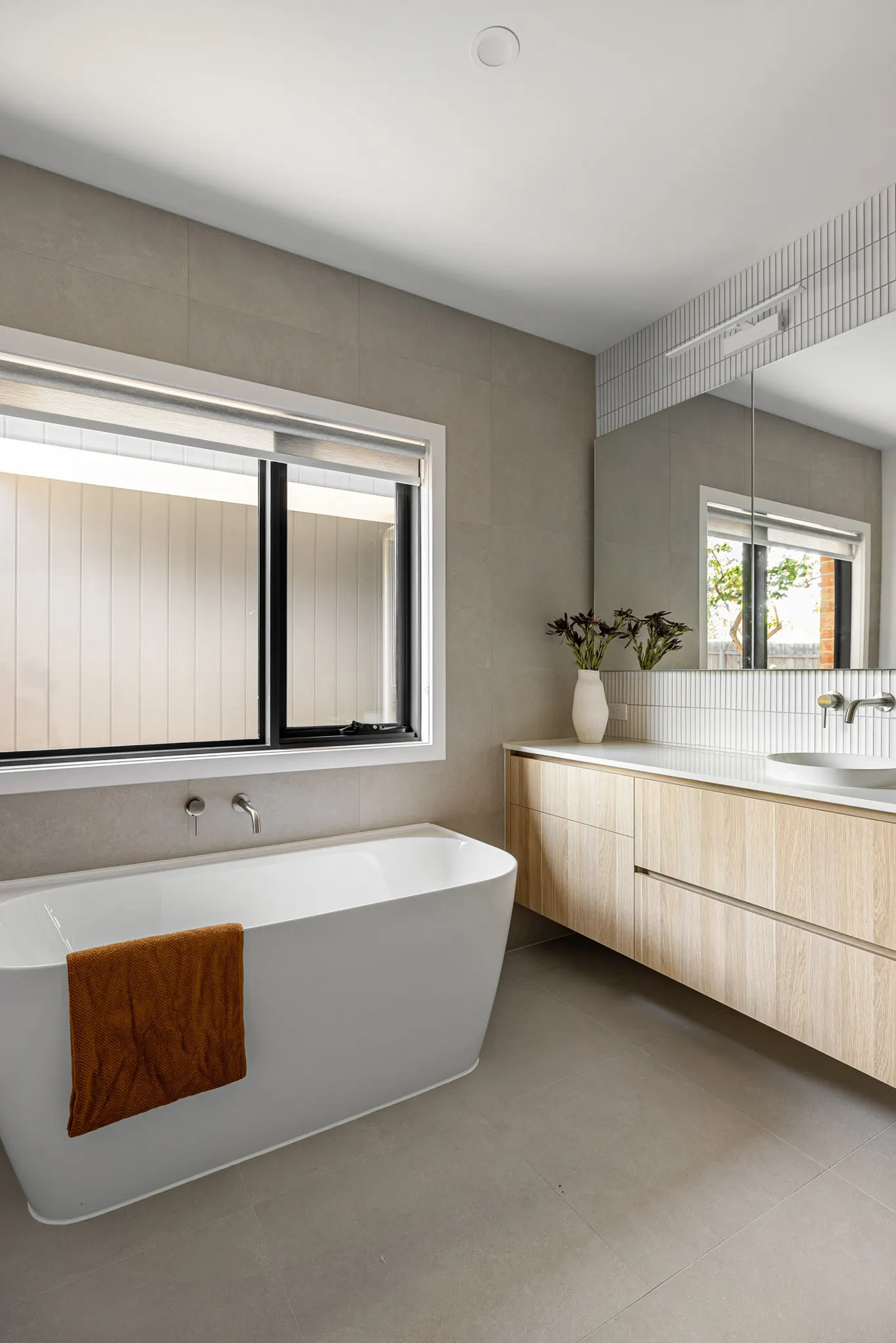 Freestanding bath beneath a window, timber vanity and large-format stone-look tiles