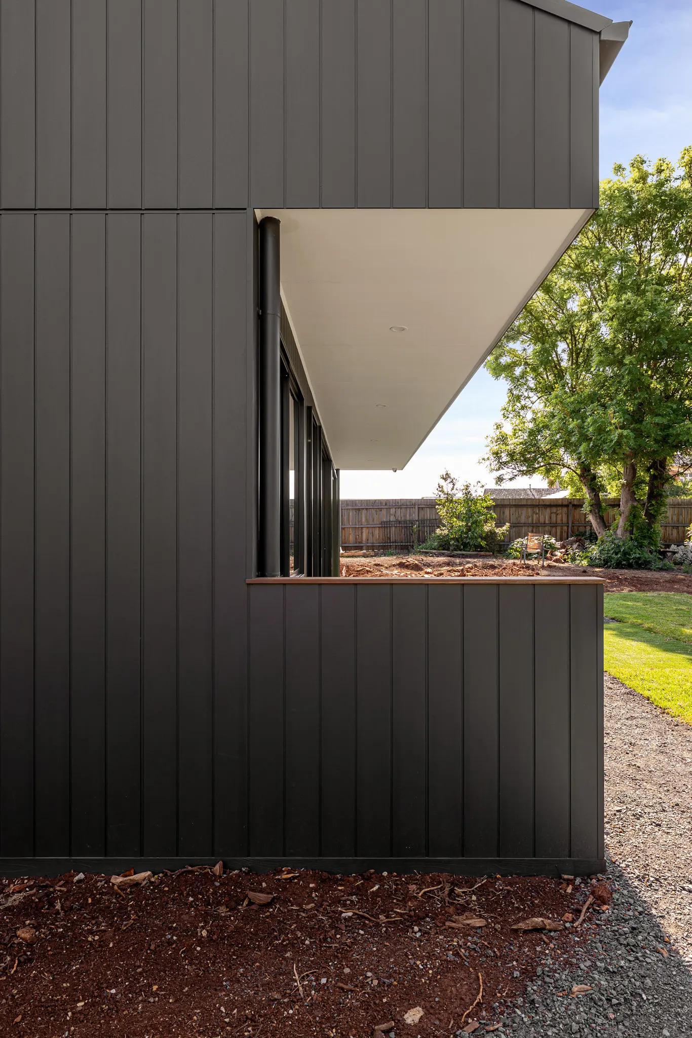 Cantilevered eave shading a window set in dark vertical cladding, garden beyond