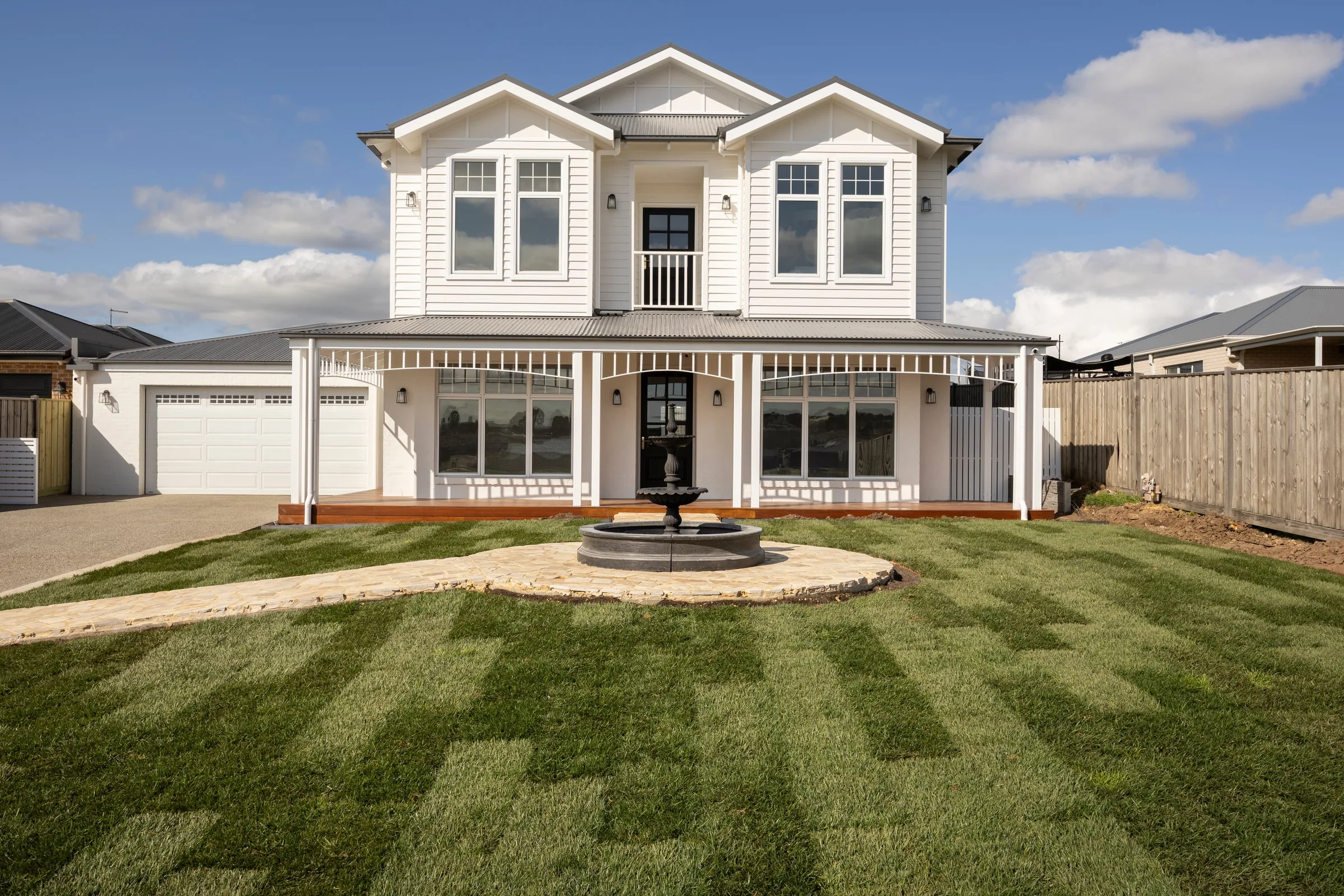 White two-storey weatherboard home with a verandah, behind a tiered fountain and new lawn