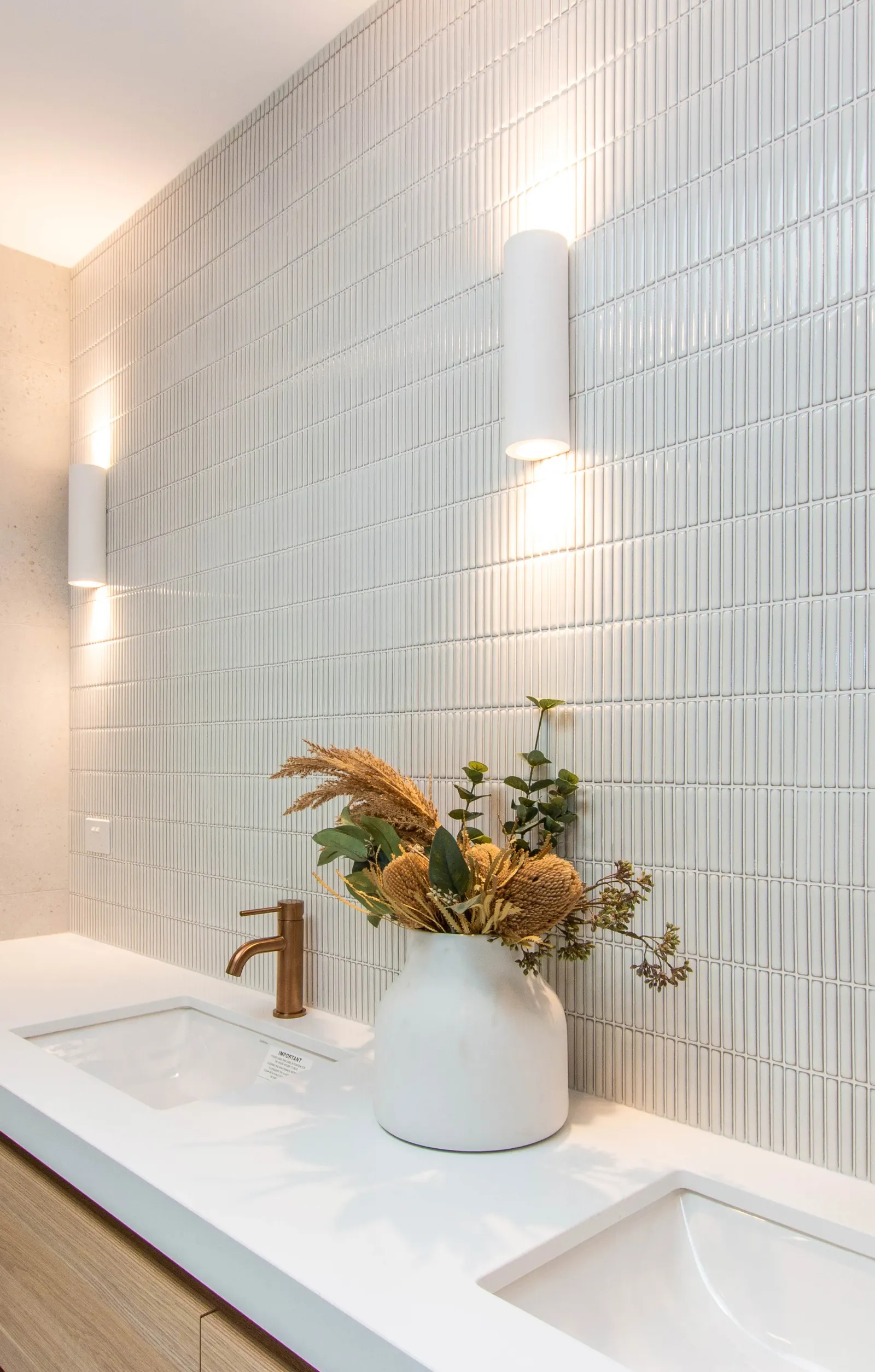 White fluted tile wall with cylindrical sconces above a stone vanity and brass tapware