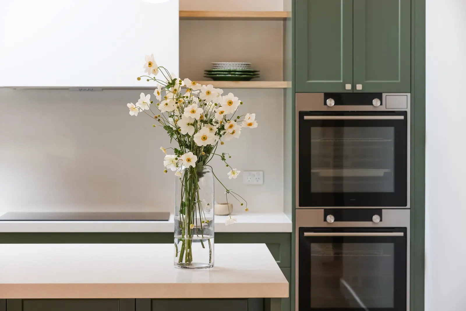 Stacked stainless wall ovens set into green shaker joinery beside timber open shelving