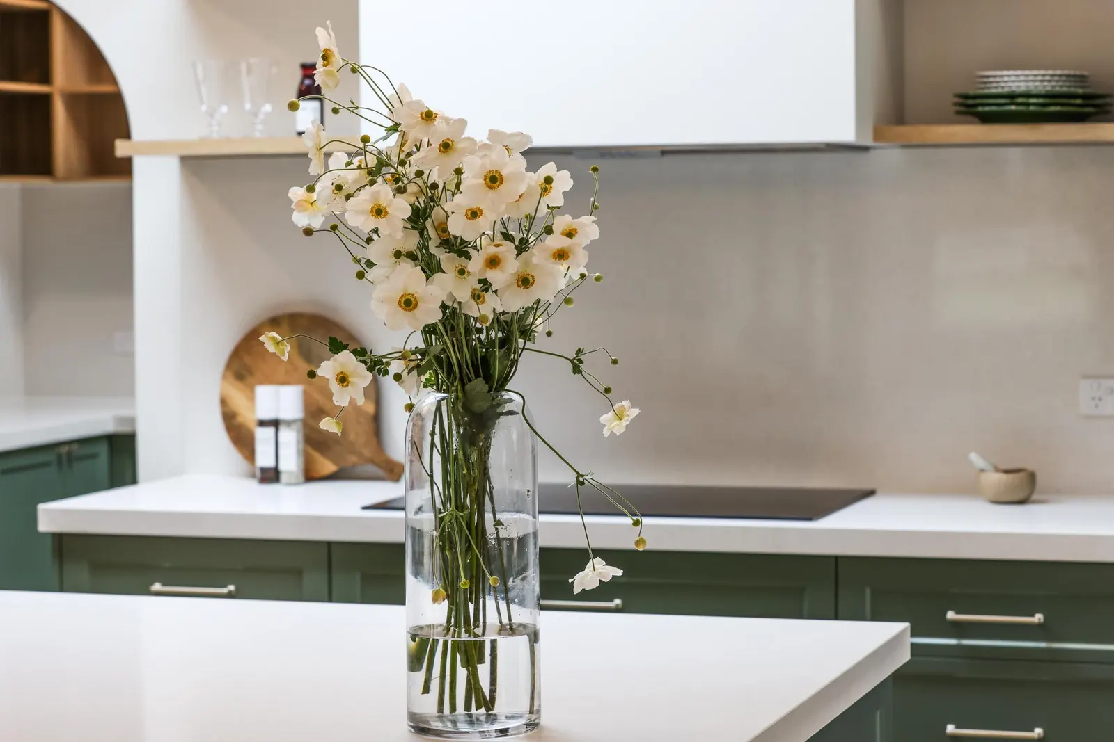 Glass vase of white blooms on the island, with green joinery and a cooktop beyond