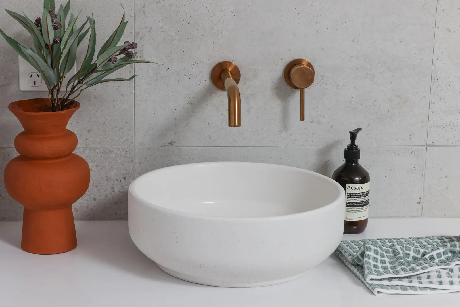 Round vessel basin with a brass wall mixer, terracotta vase and concrete-look tiling