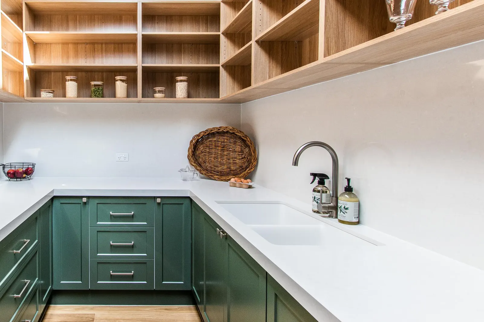 Green shaker joinery and white stone benchtops beneath timber shelving in the pantry
