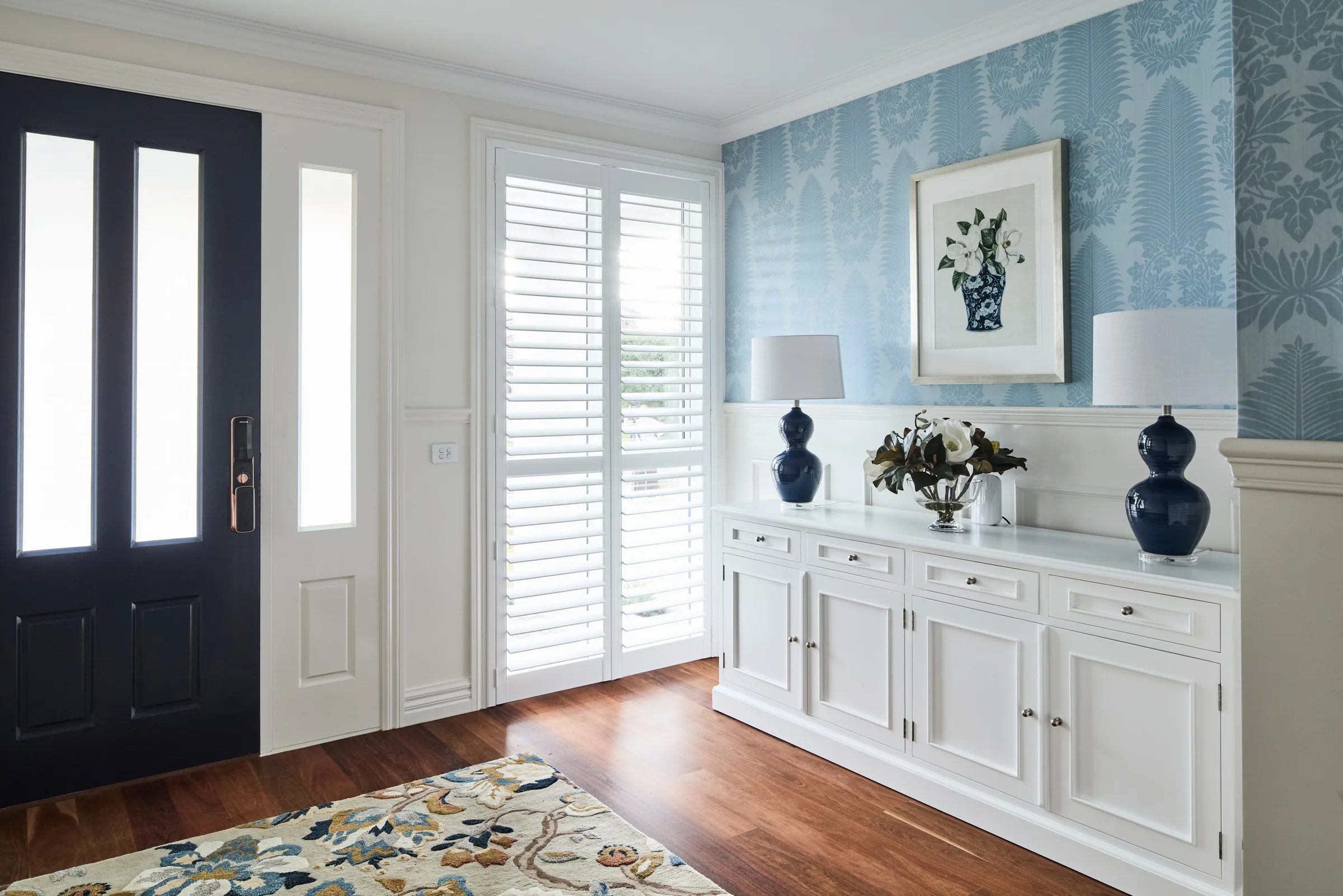 Front door and plantation shutters beside the sideboard, blue wallpaper above panelled walls