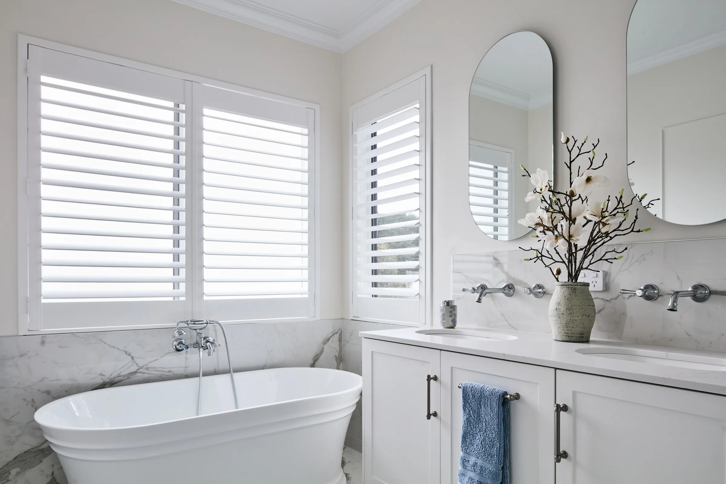 Freestanding bath beneath plantation shutters in the ensuite, arched mirrors over a twin vanity