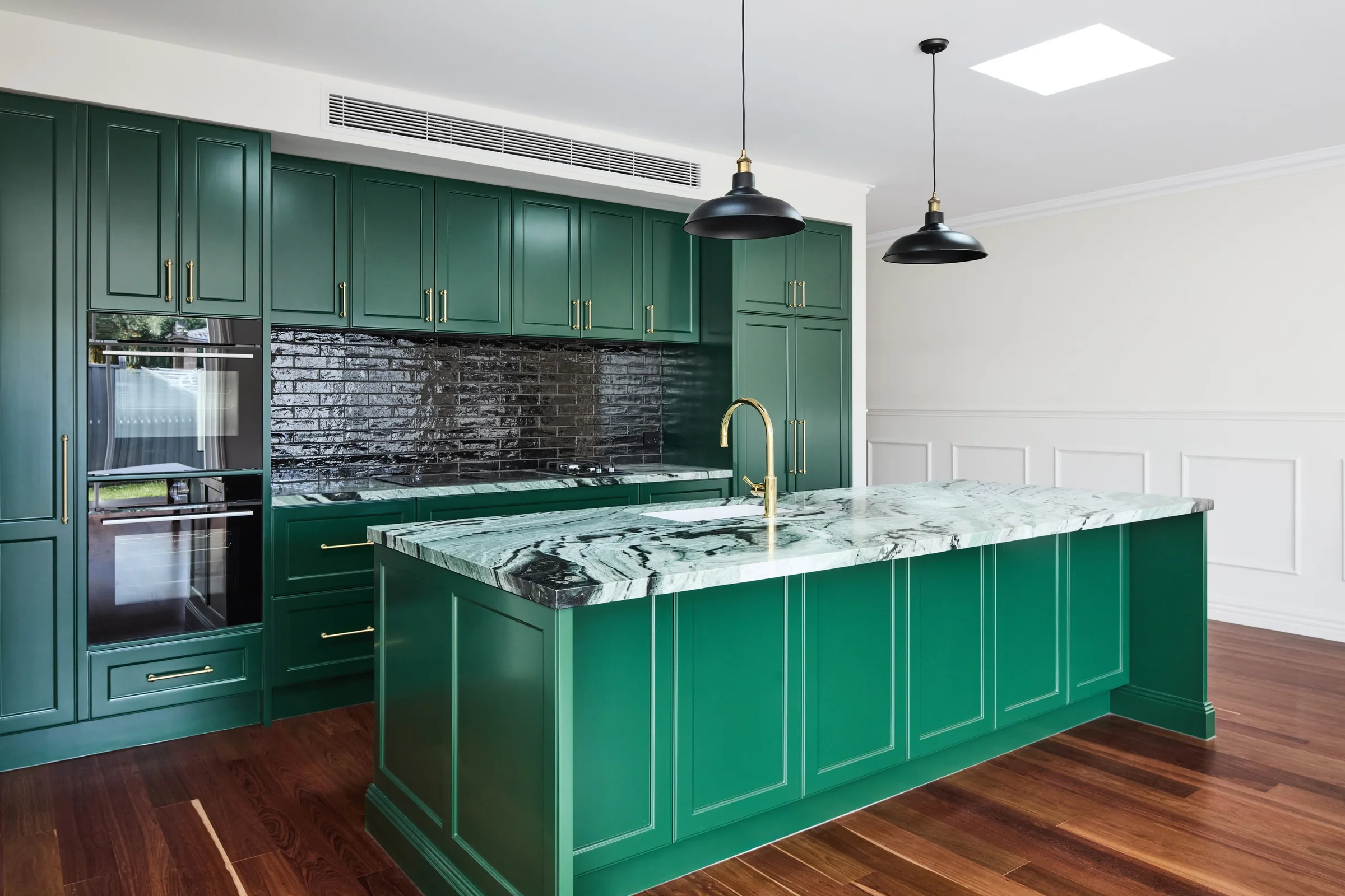Green kitchen from the side, with wall ovens, an island cooktop and brass tapware