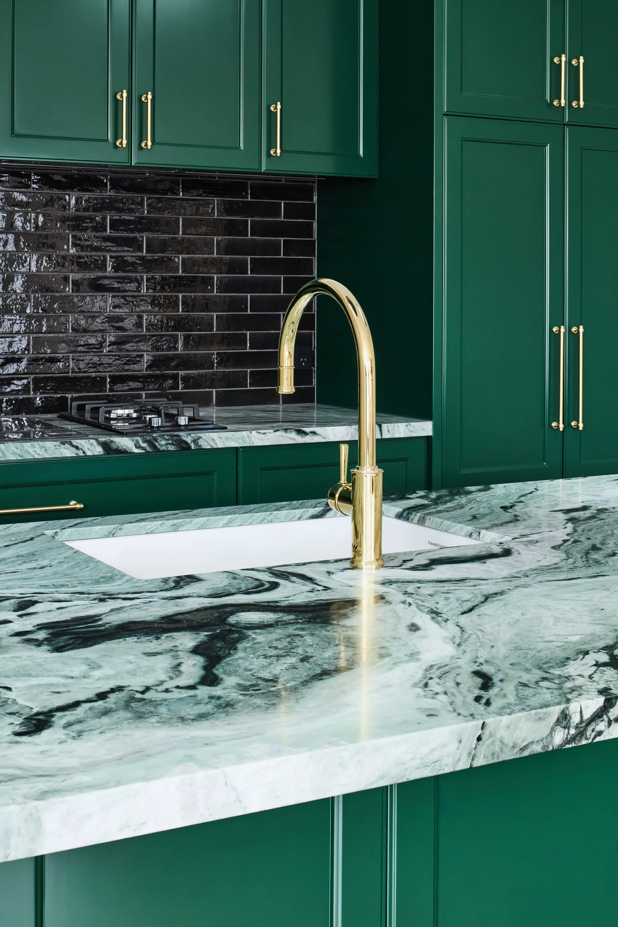 Brass gooseneck mixer over an undermount sink in a green marble benchtop, black brick splashback