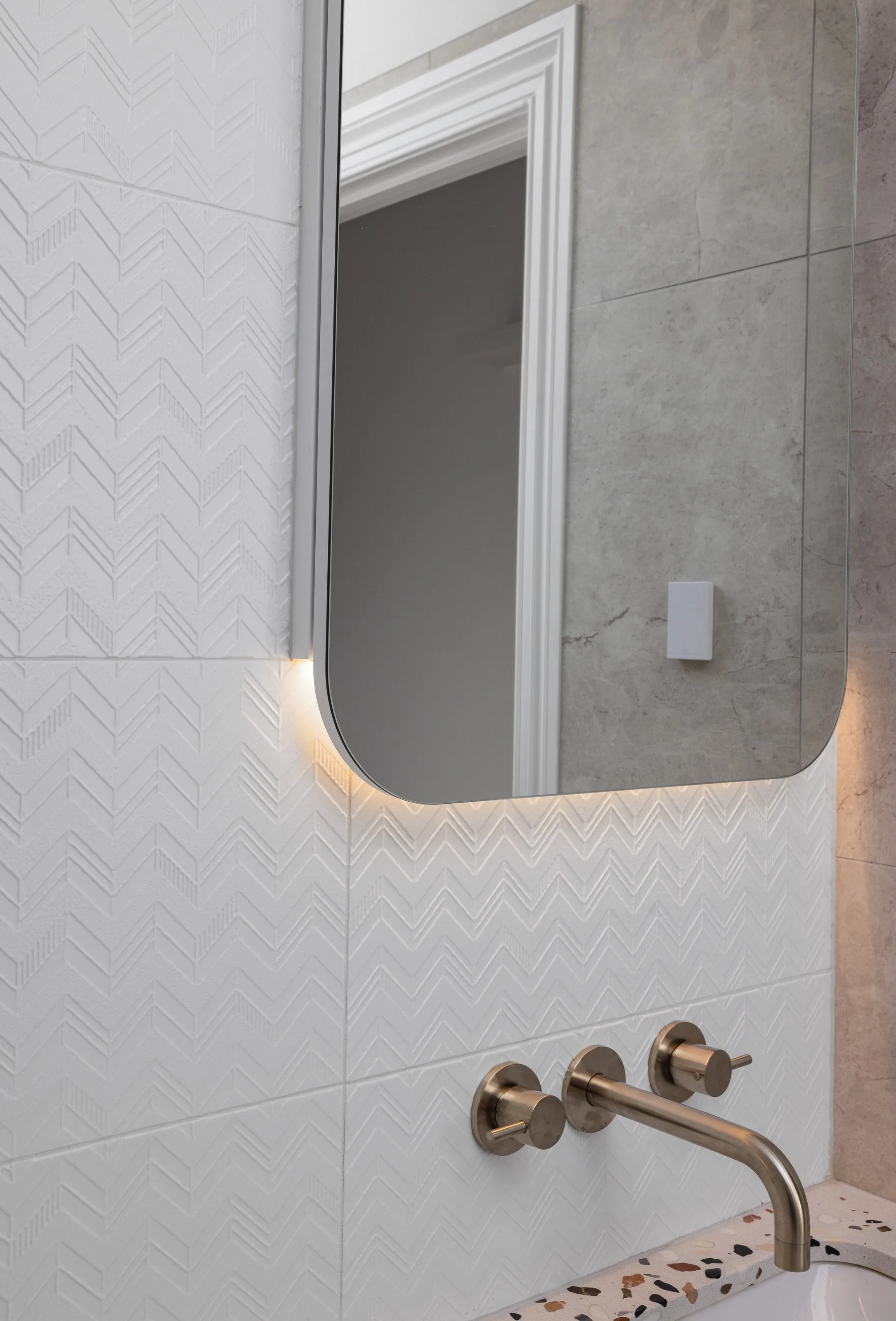 Backlit rounded mirror above brushed nickel wall tapware, chevron-textured white tiles and terrazzo benchtop