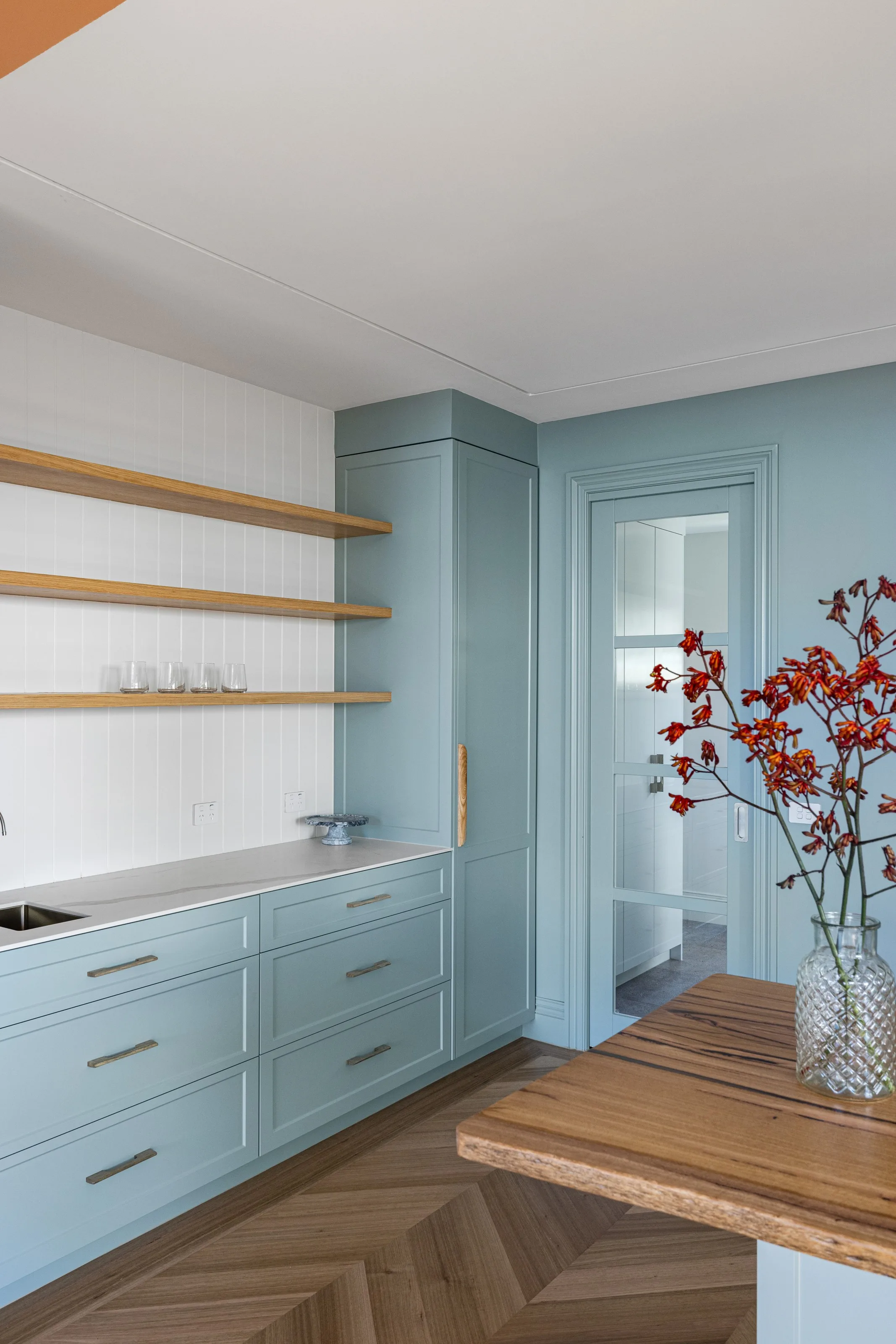 Pale blue shaker joinery with oak floating shelves, white tiled splashback and a doorway to the pantry