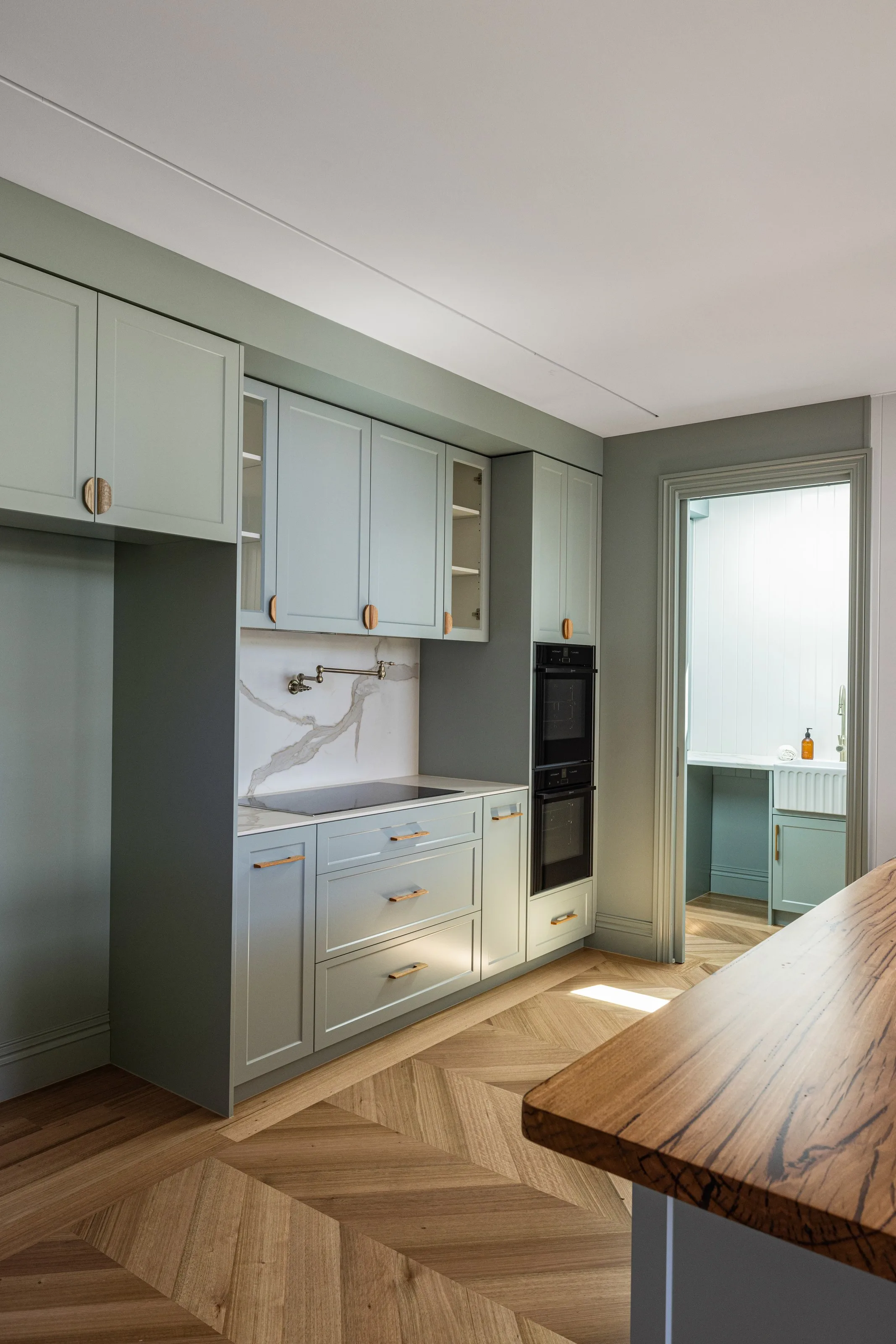 Sage kitchen joinery with a veined stone splashback, induction cooktop and black wall ovens