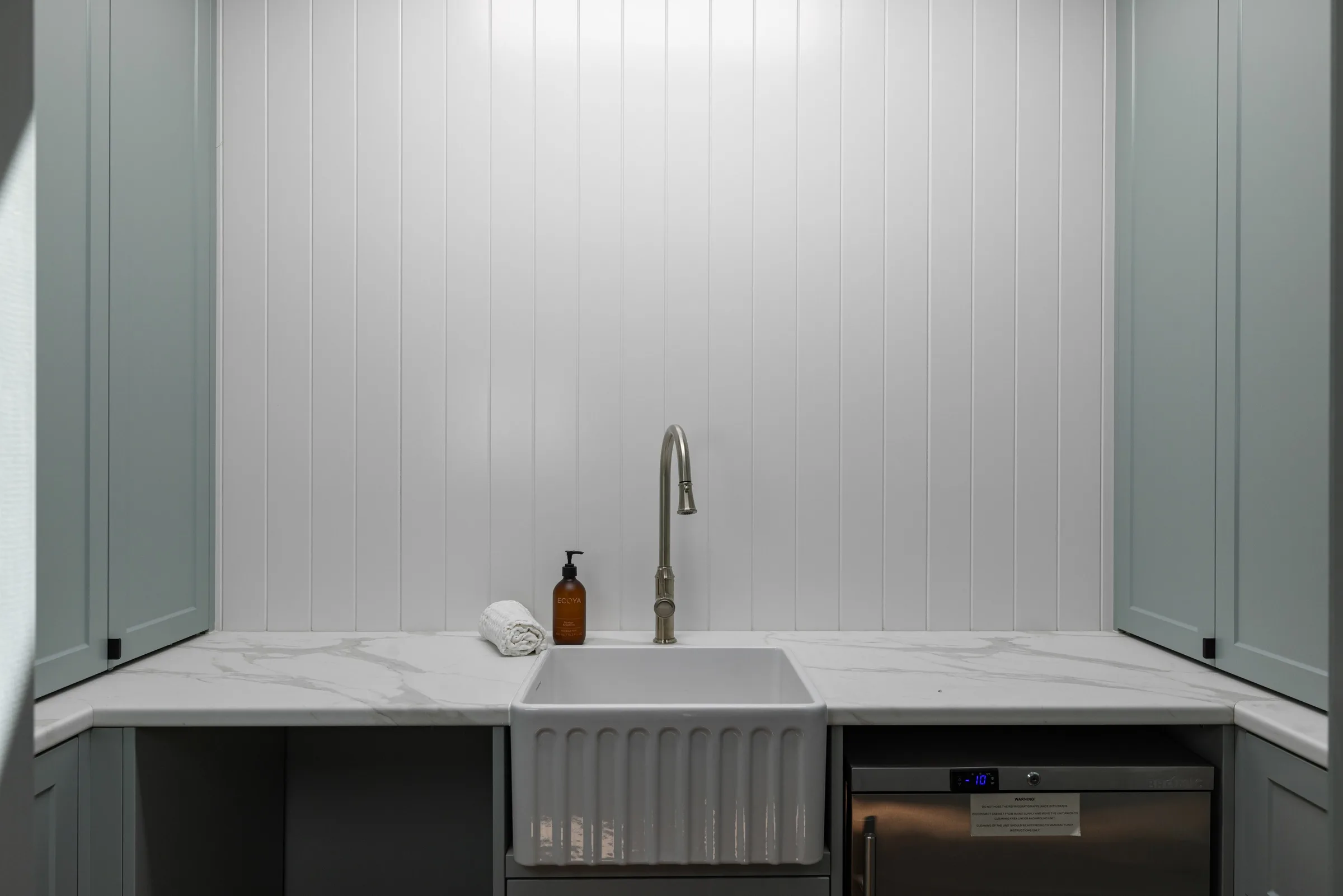 Fluted apron sink centred on a stone benchtop against white VJ panelling, dishwasher alongside