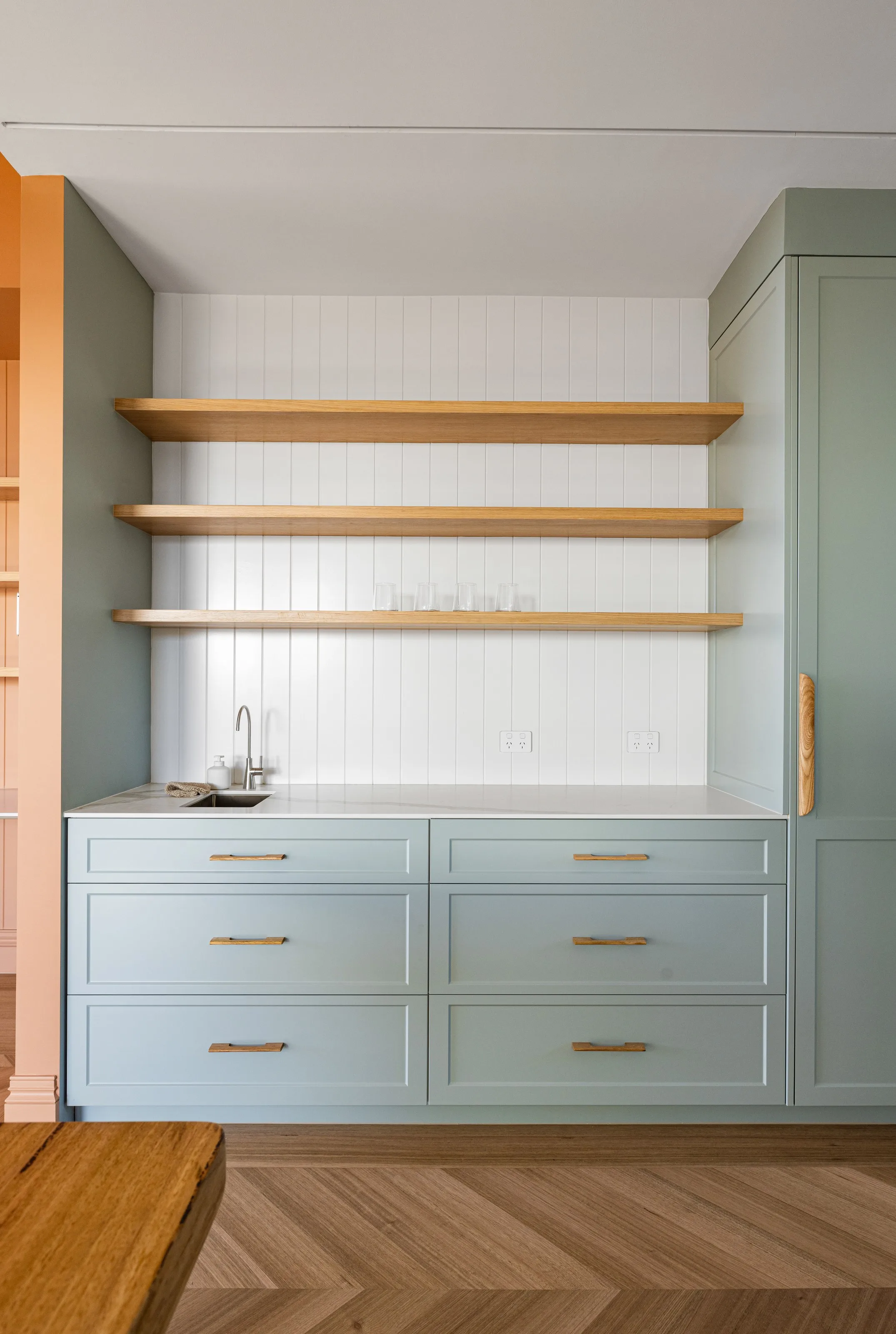 Pale green joinery with oak floating shelves, white VJ splashback and a small bar sink
