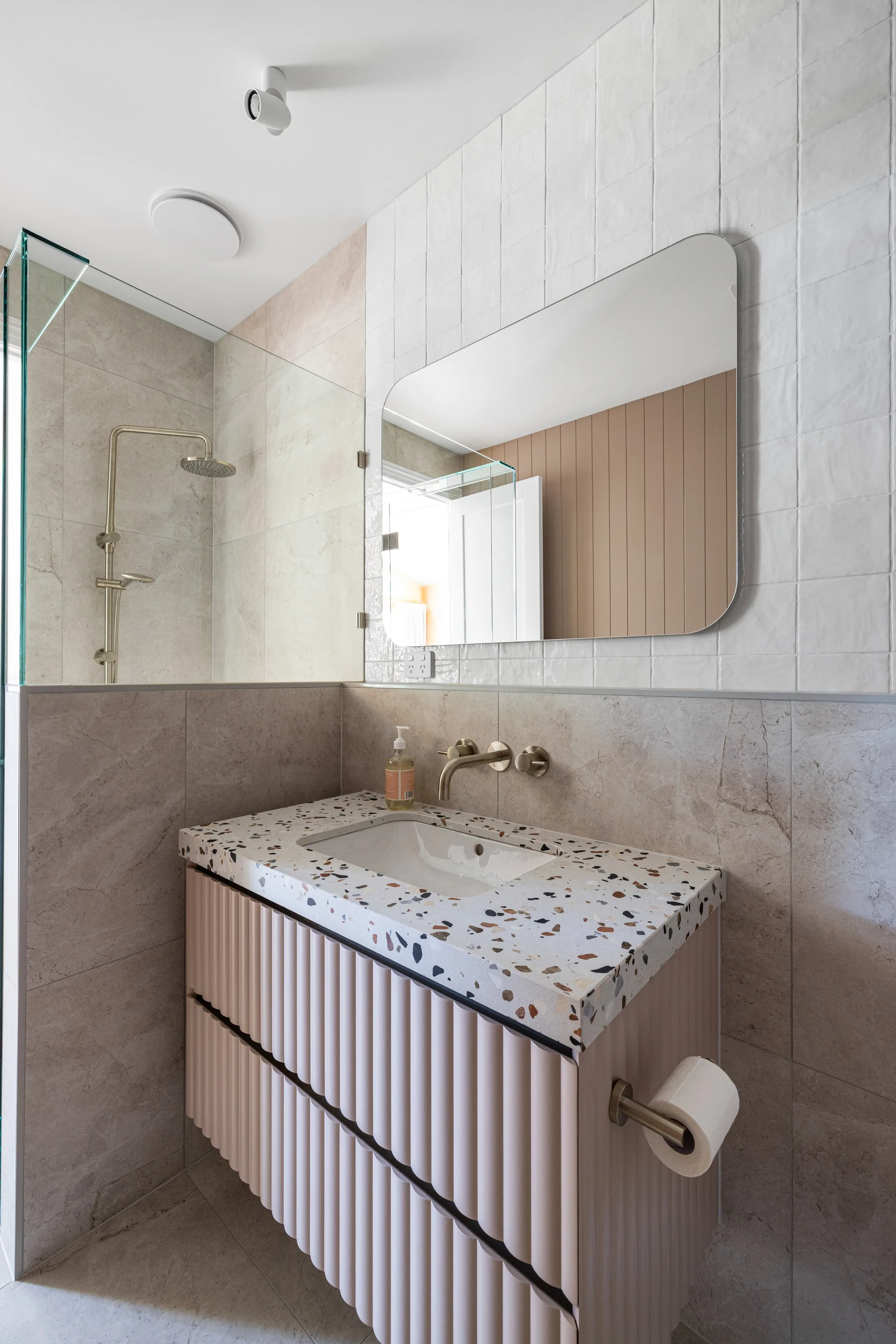 Fluted blush vanity with terrazzo top and wall tapware, walk-in shower behind glass