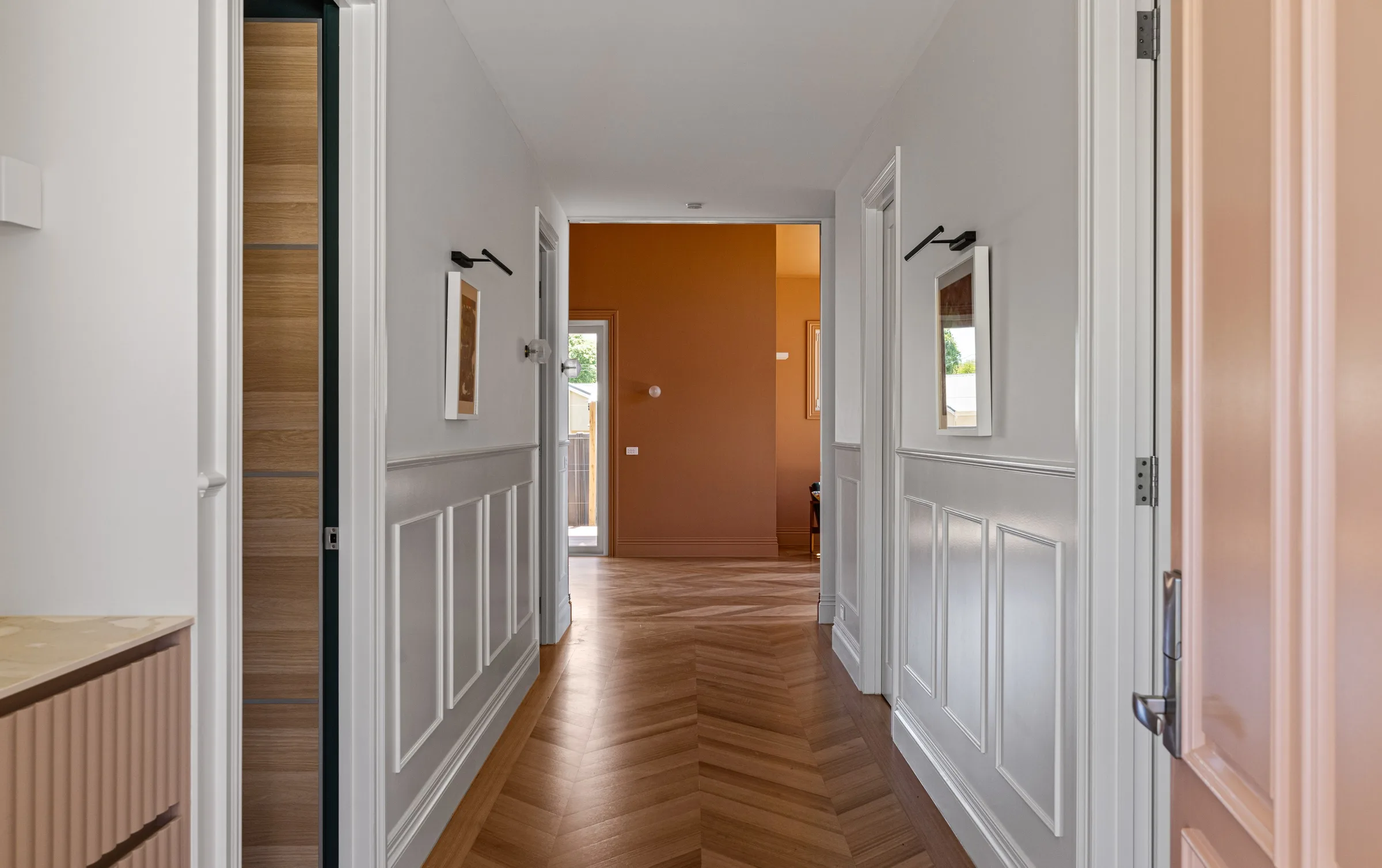 Hallway with white wainscoting and chevron parquet leading to a terracotta room beyond