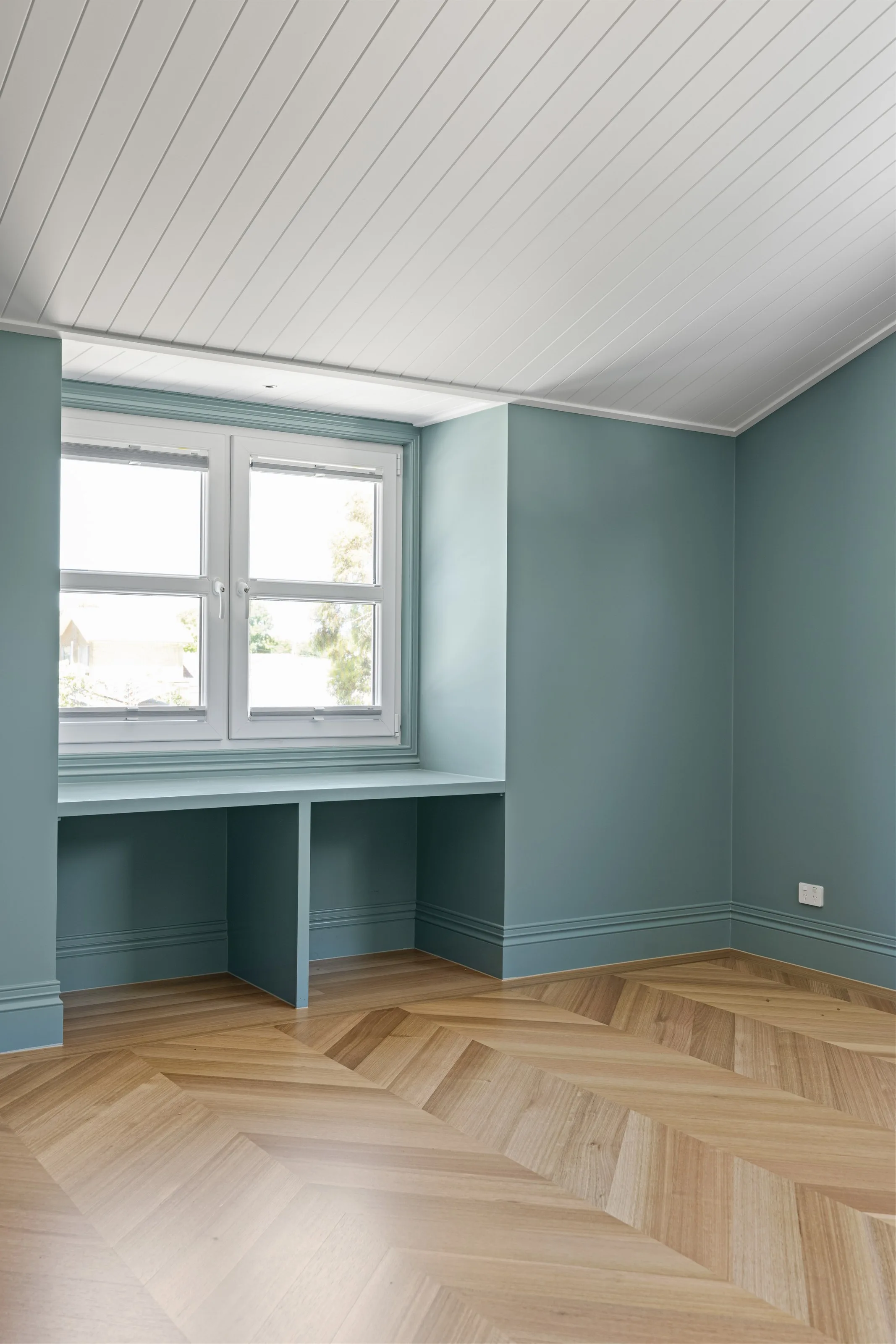 Study nook with a built-in desk under the window, blue-green walls and a white lined ceiling