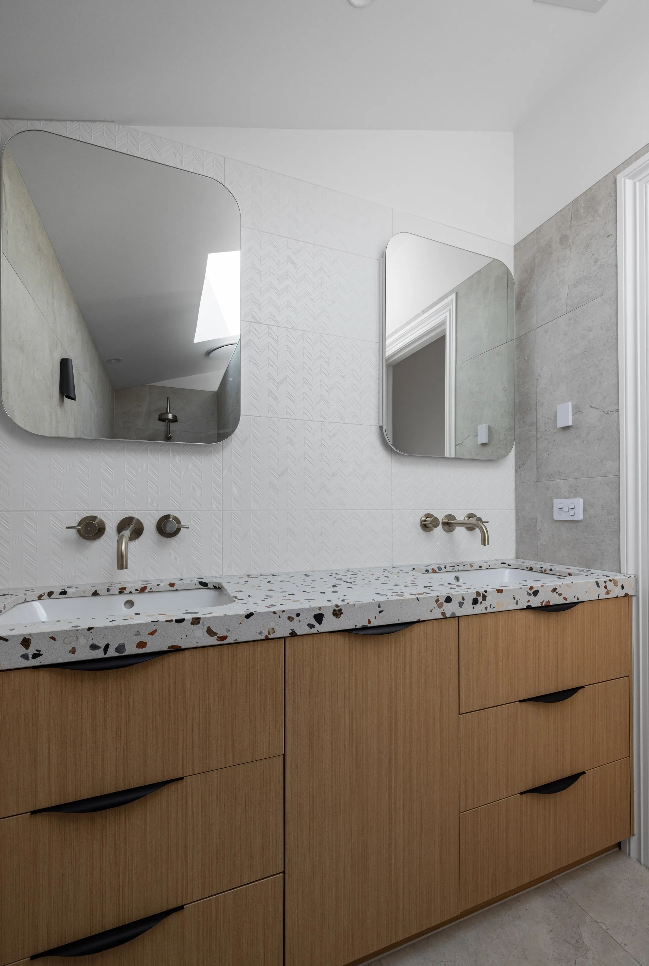 Twin basins set into terrazzo above timber joinery, against chevron-textured white tiles
