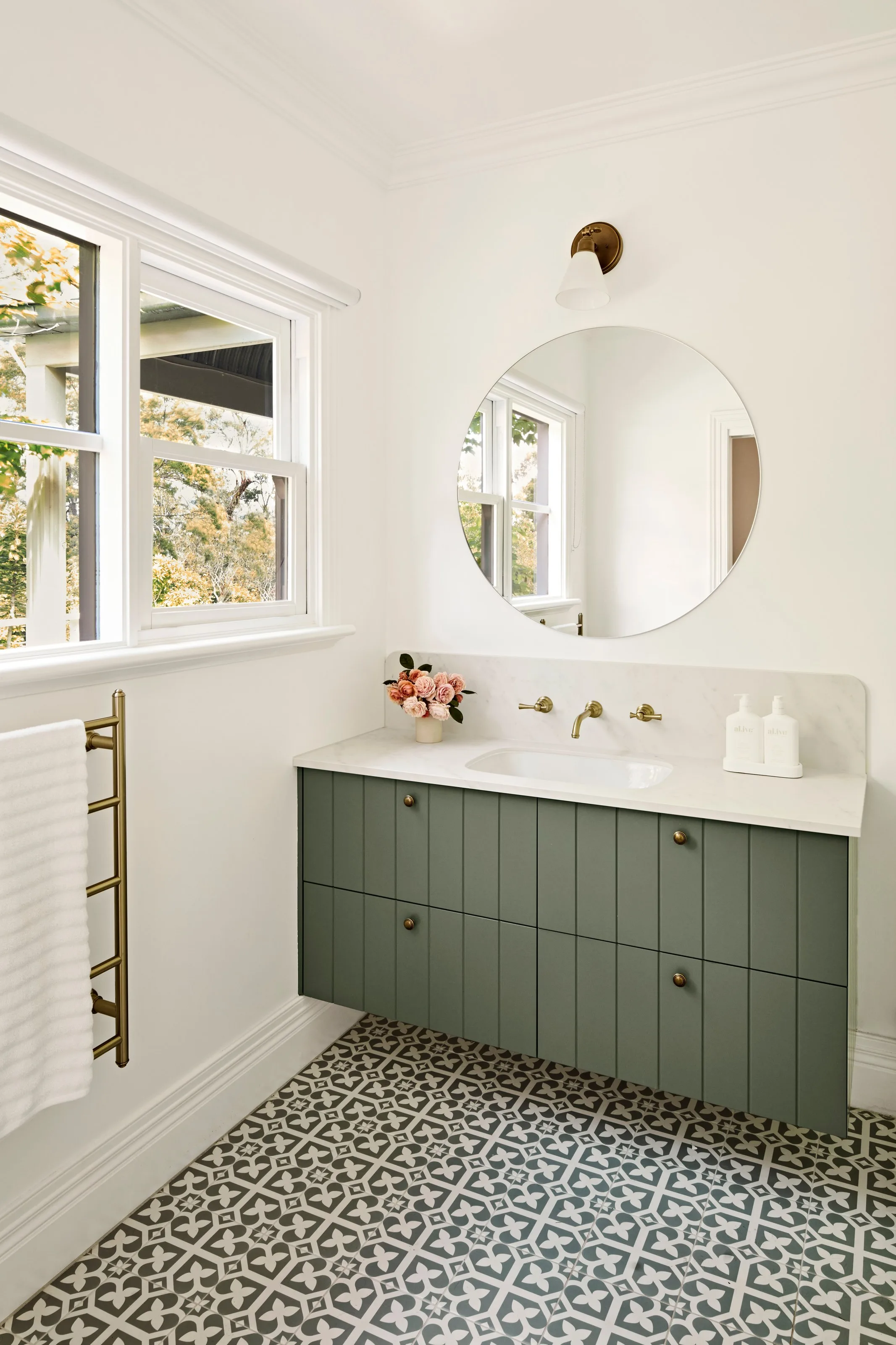 Sage green vanity with stone benchtop, round mirror and brass tapware over patterned floor tiles