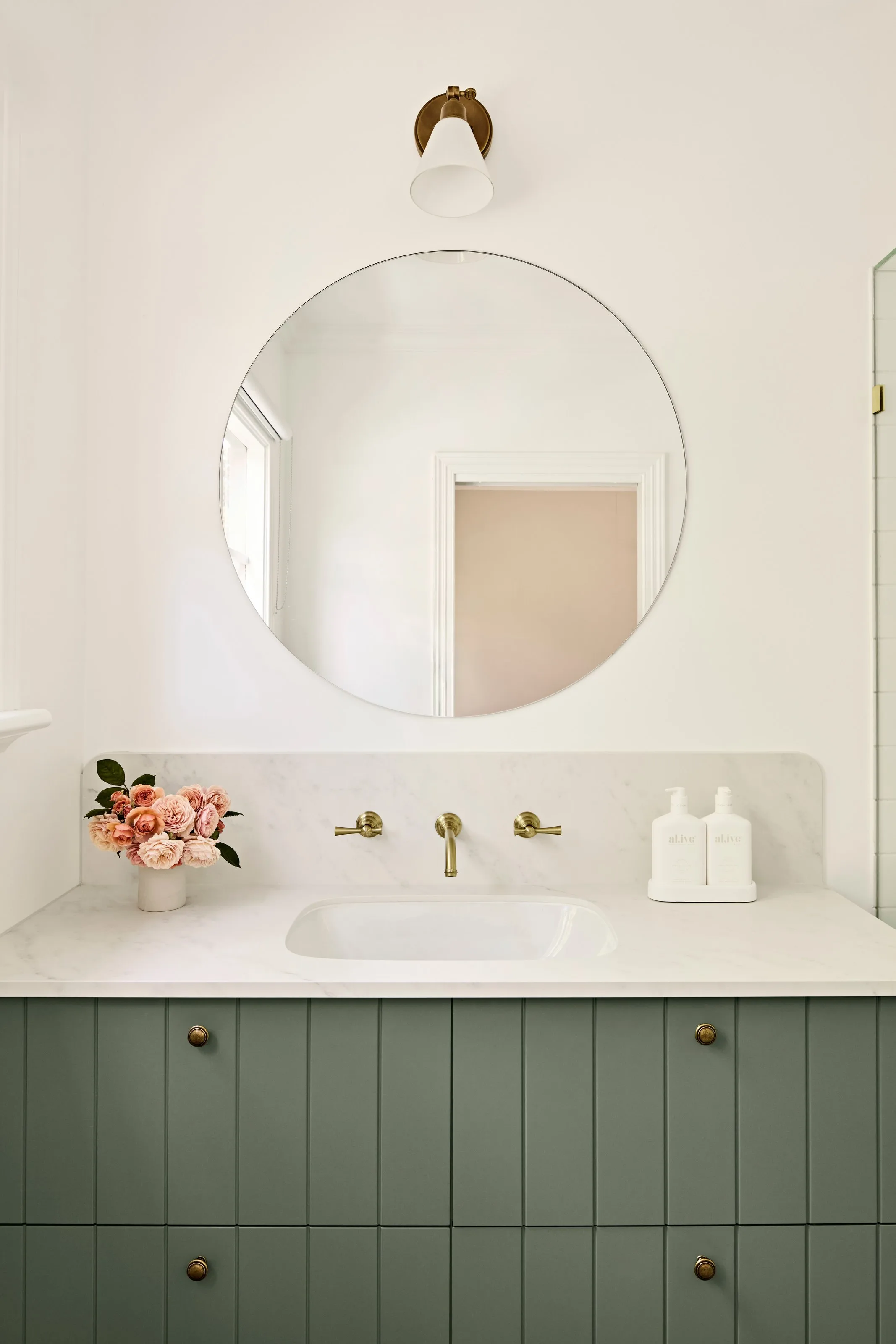 Round mirror and brass wall light above a stone benchtop with wall-mounted tapware