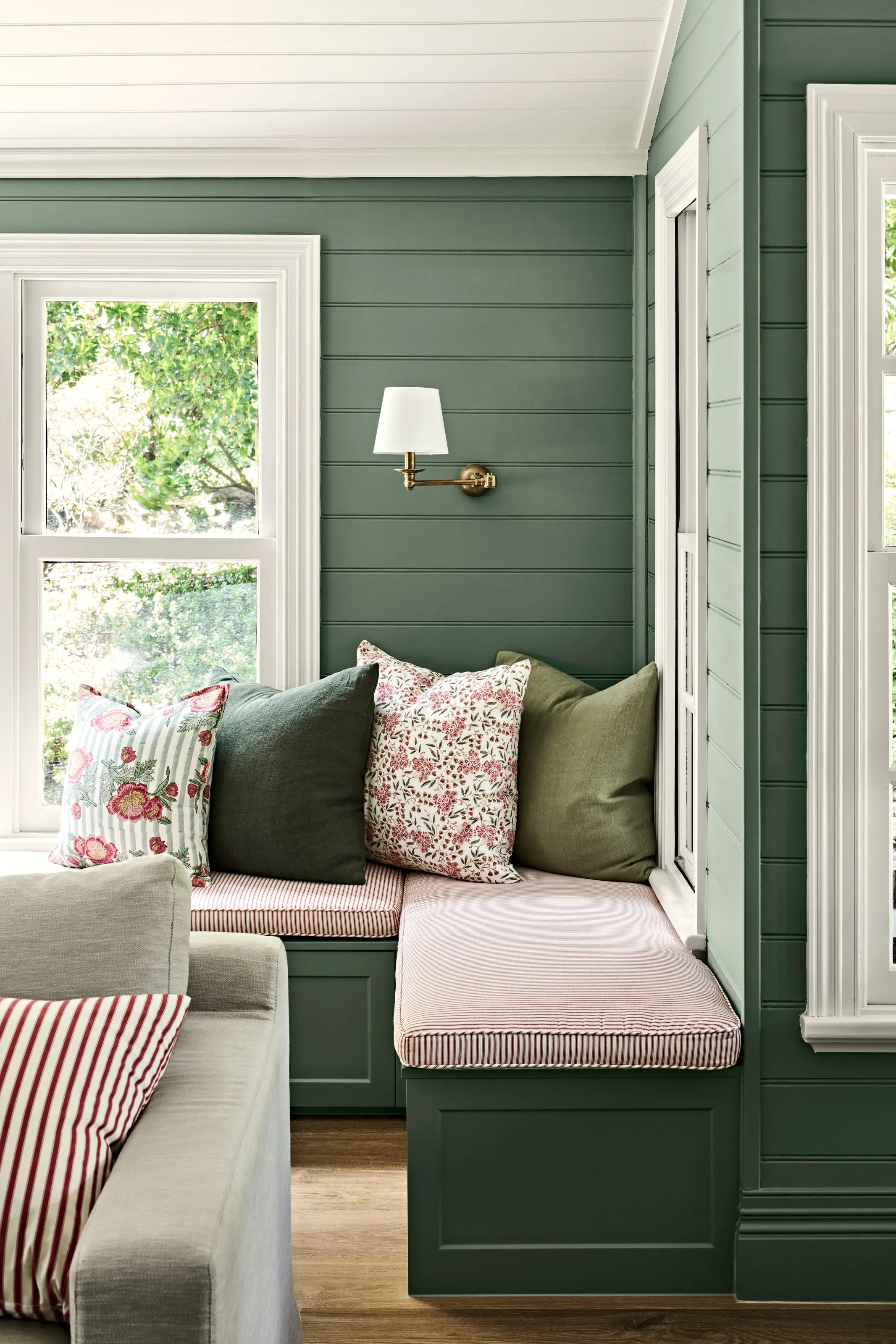 Corner window seat with piped pink cushions under a brass swing-arm light