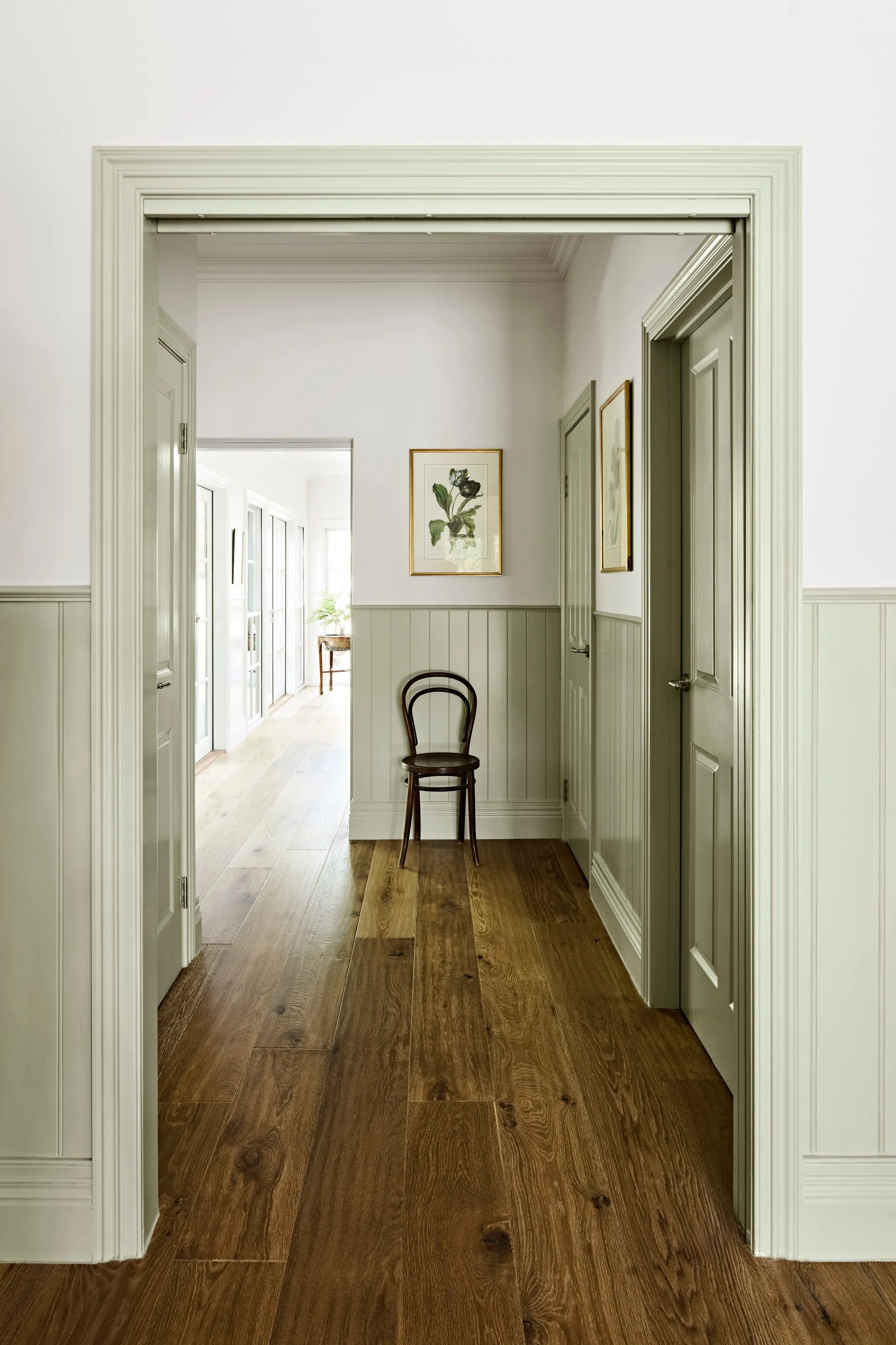 Hallway with sage wainscoting, panelled doors and a bentwood chair on dark boards