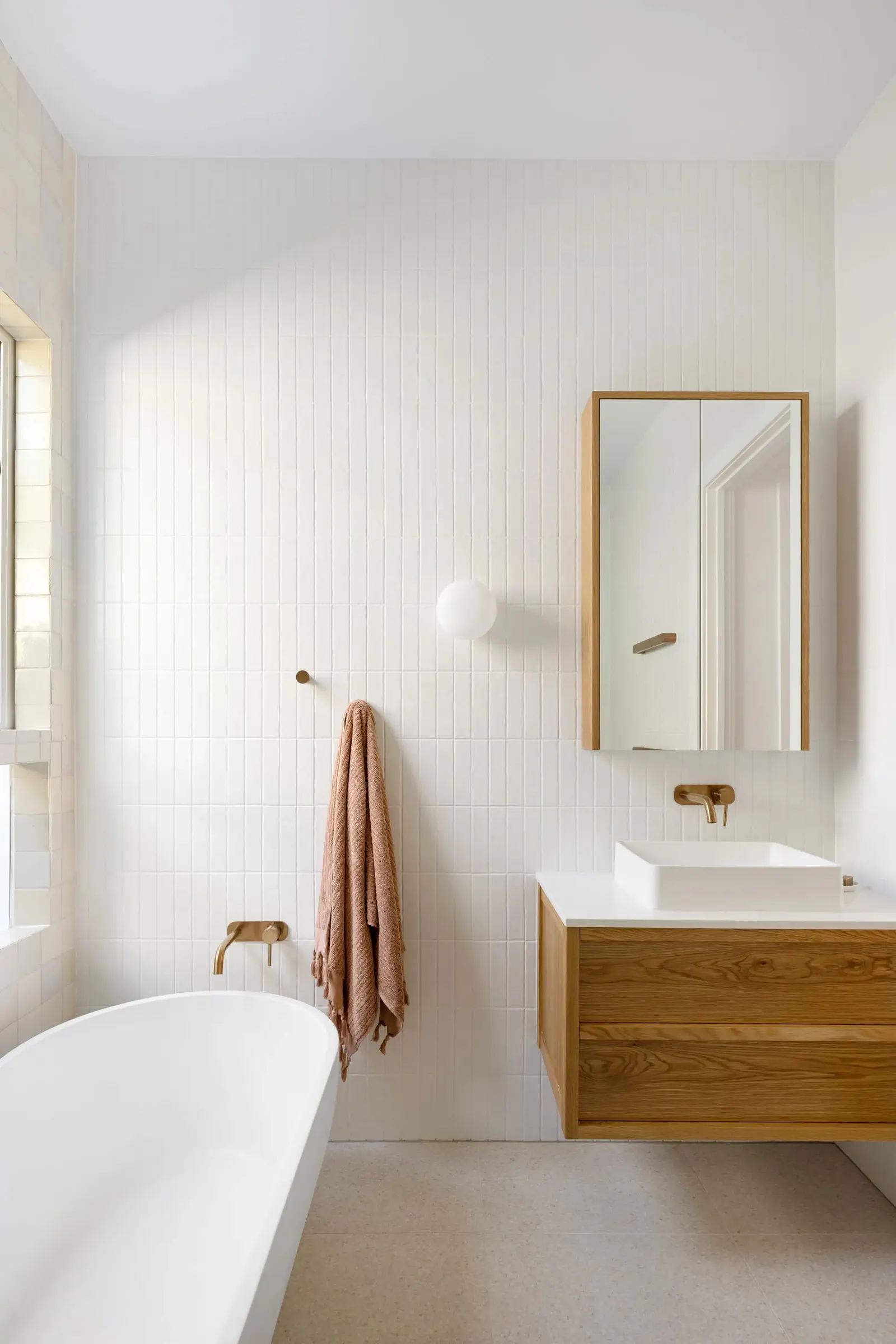 Oak-framed mirror cabinet and globe wall light above a wall-hung vanity, with a towel on a brass hook beside the bath