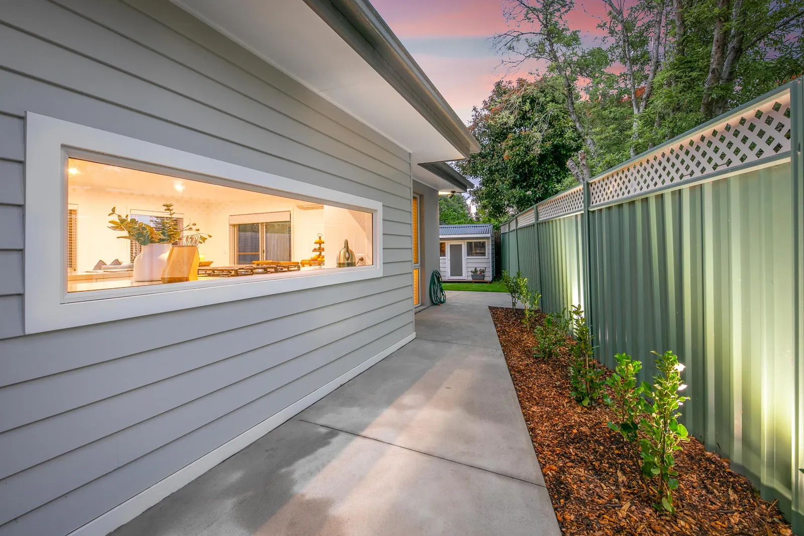 Side path beside weatherboard cladding, a long picture window glowing from the lit kitchen