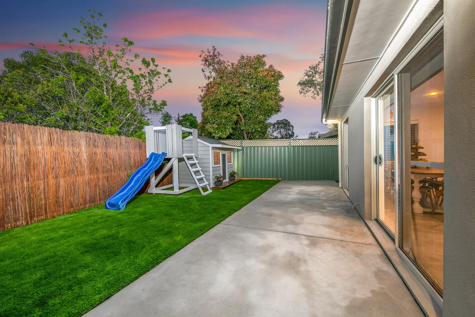Rear yard at dusk with lawn, a children&rsquo;s cubby and slide, and sliding doors to the living area