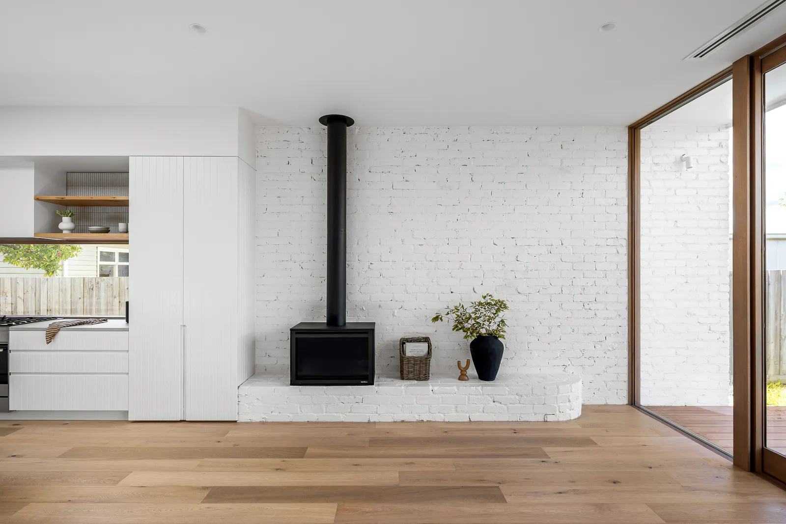 Black wood heater on a white painted brick wall, timber floors and kitchen joinery beyond