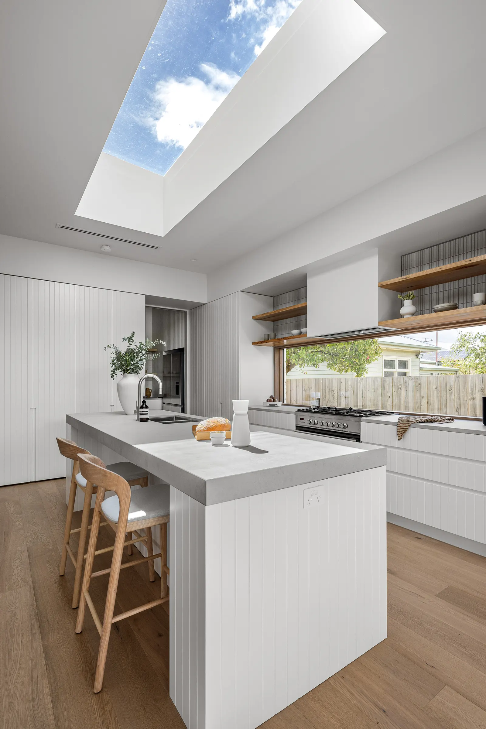Kitchen island with stone benchtop under a large skylight, oak stools and fluted white joinery