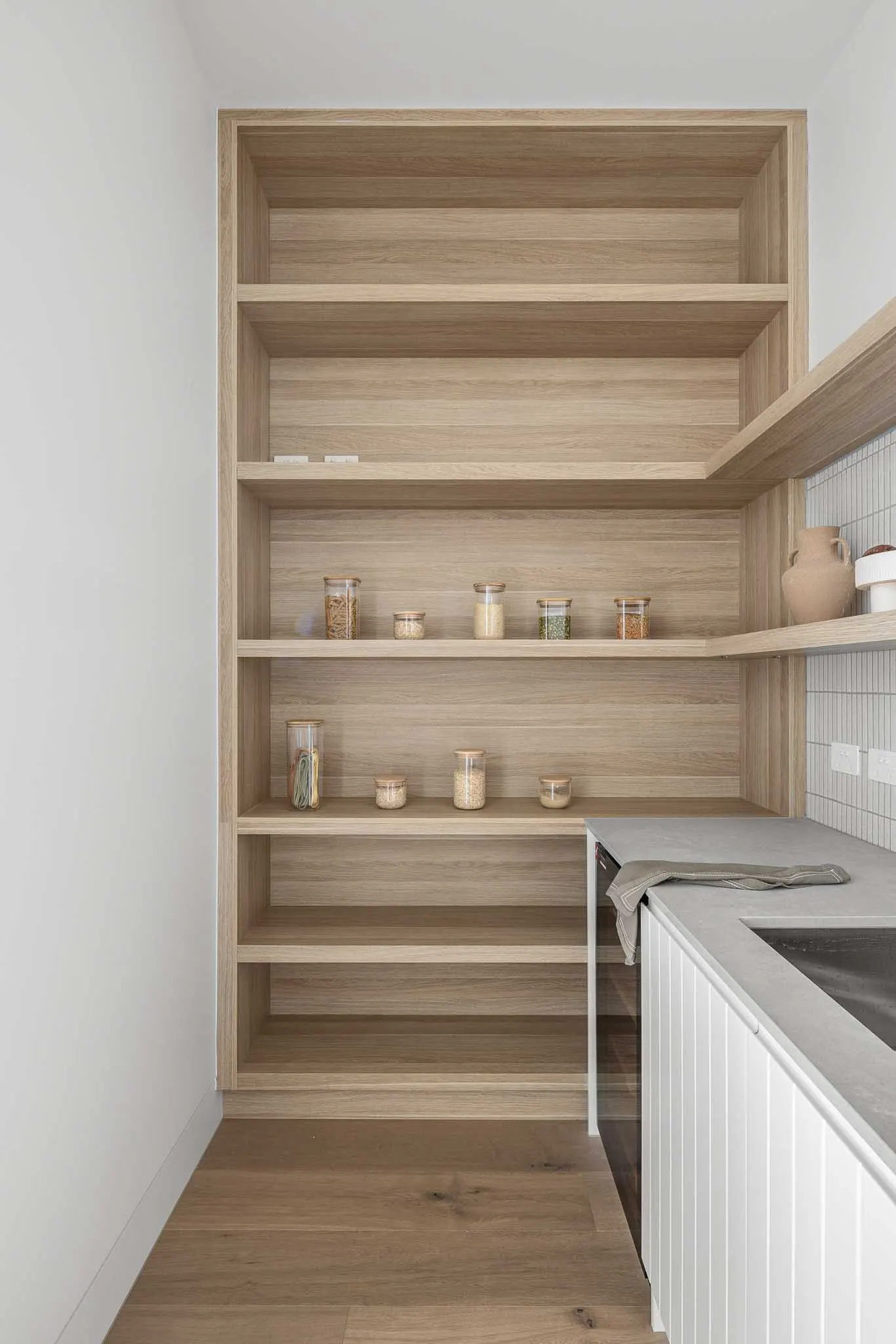 Walk-in pantry lined with oak shelving beside a stone benchtop and white tiled wall
