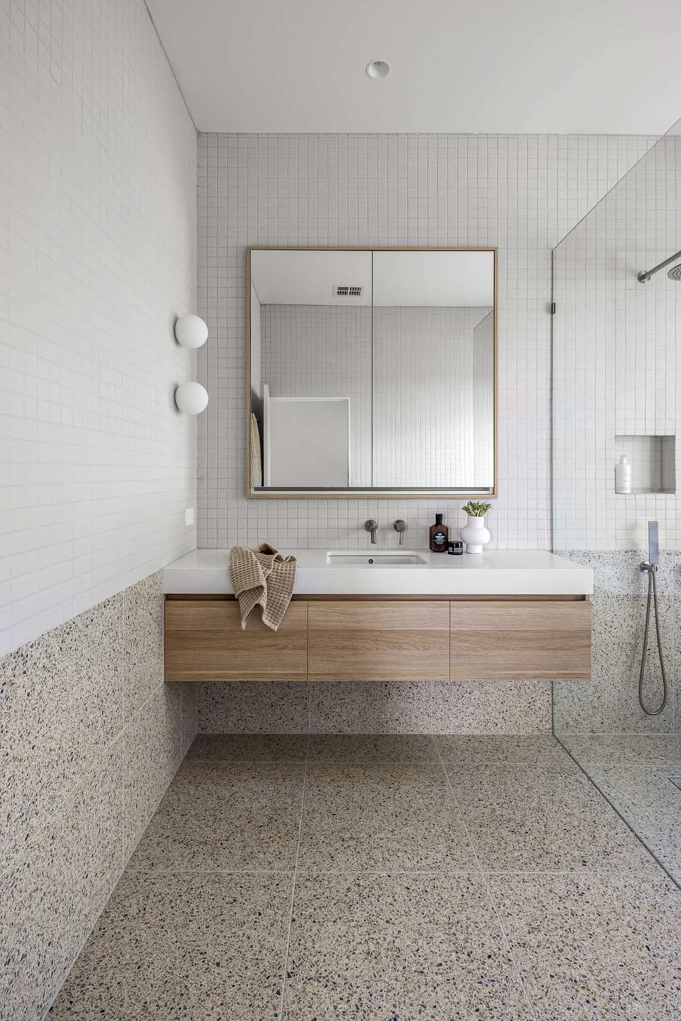 Bathroom in white mosaic tile, oak floating vanity and terrazzo floor, shower at right