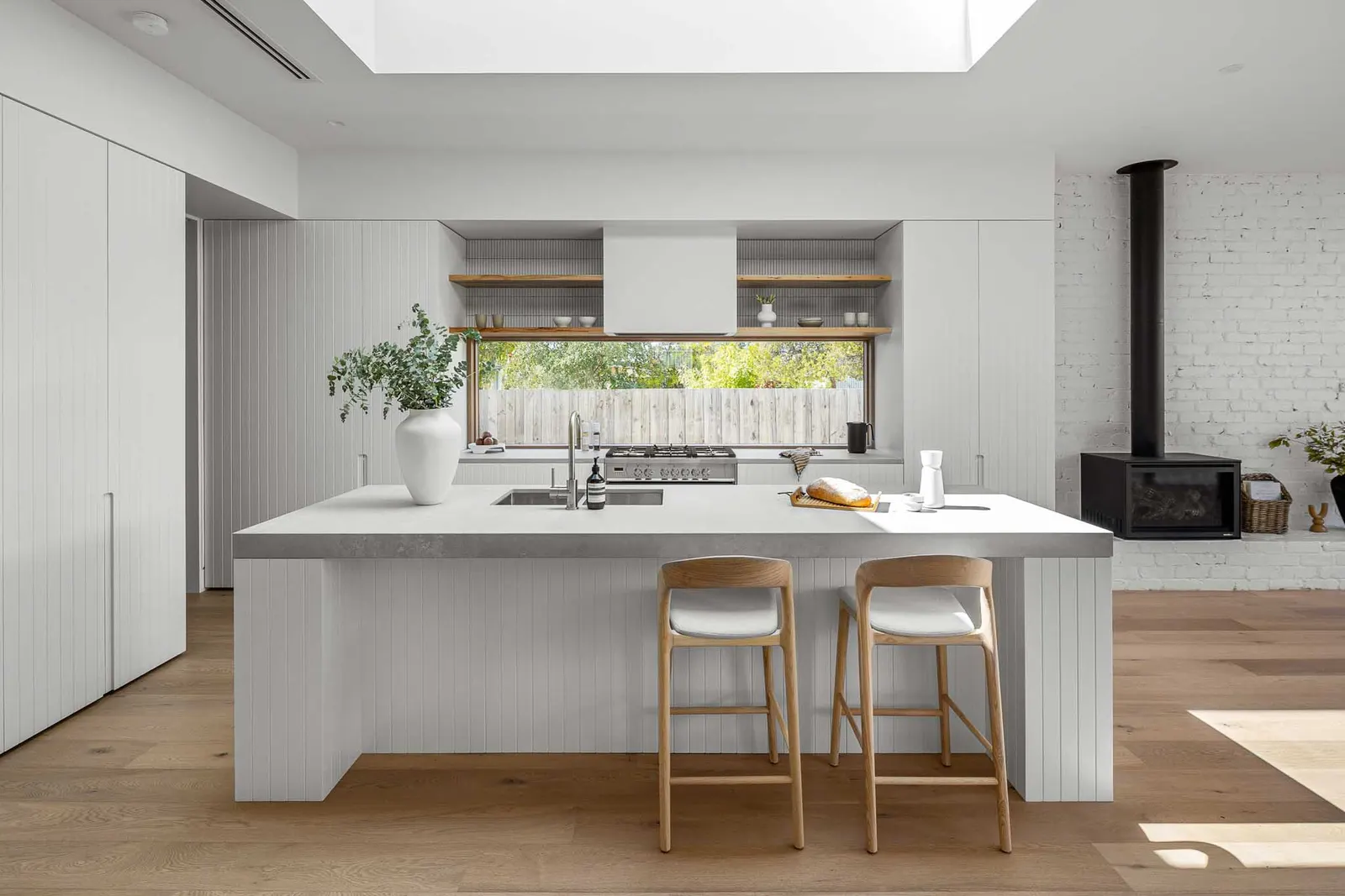 Open kitchen at Yaldwyn St, Kyneton &mdash; island bench, skylight and wood heater on white brick